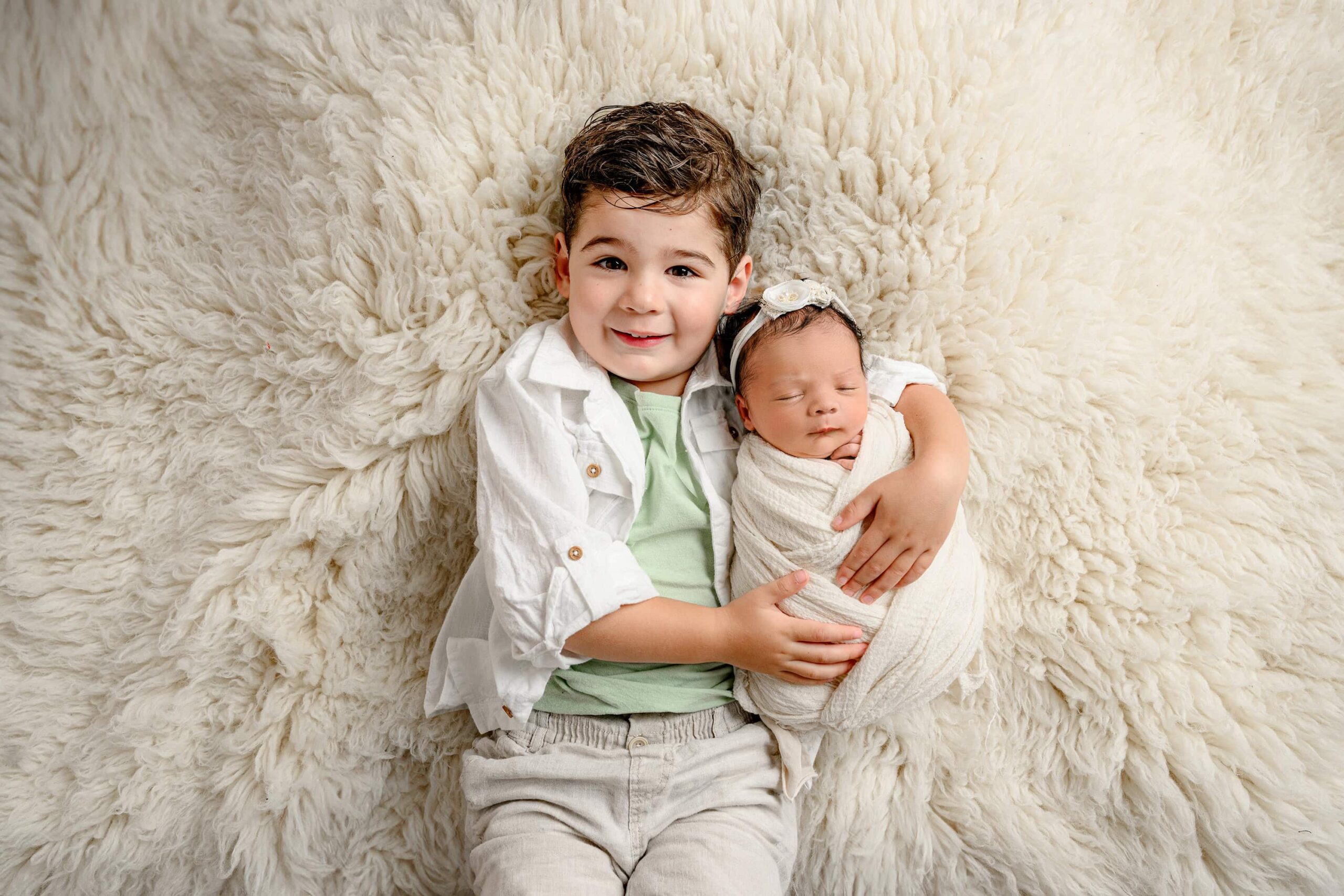 Newborn baby girl wrapped in cream is being held by her big brother while laying on a cream rug