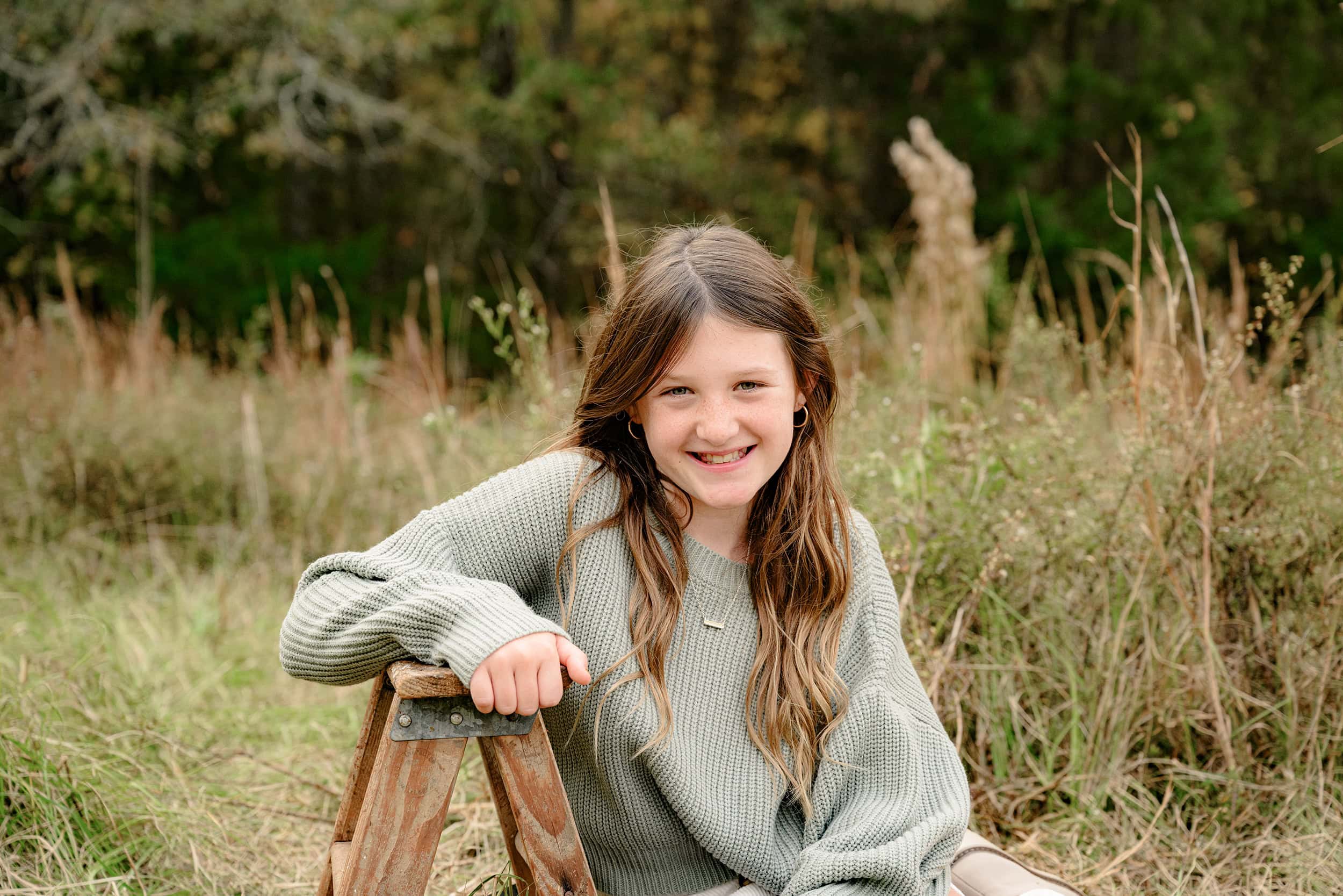girl dressed in green sweater leans on a small ladder in tall fall grasses of a charlotte area park