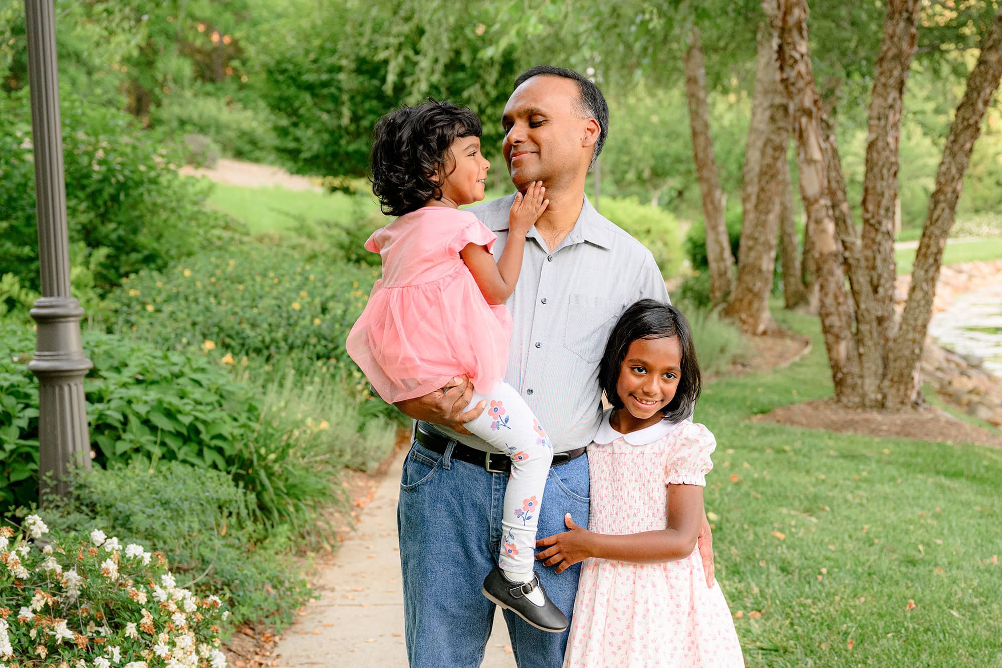 dad holds youngest daughter and has oldest daughter beside him smiling sweetly at his youngest on a summer day in a charlotte nc park]]][]][[]]