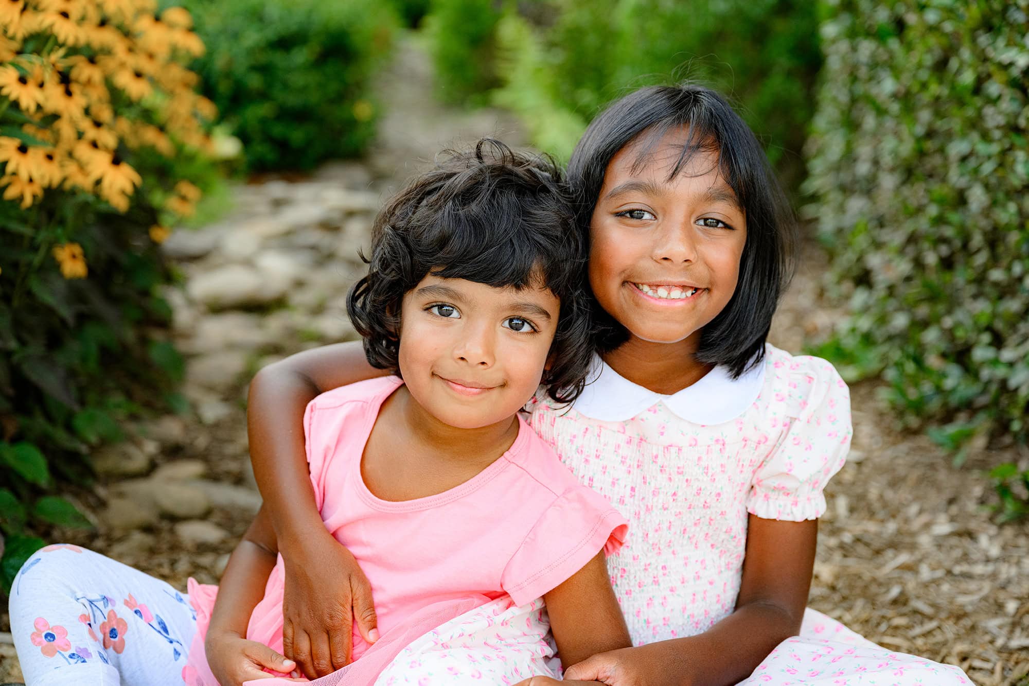 Two sisters sit together in a flower garden on a warm summer day in charlotte with their family
