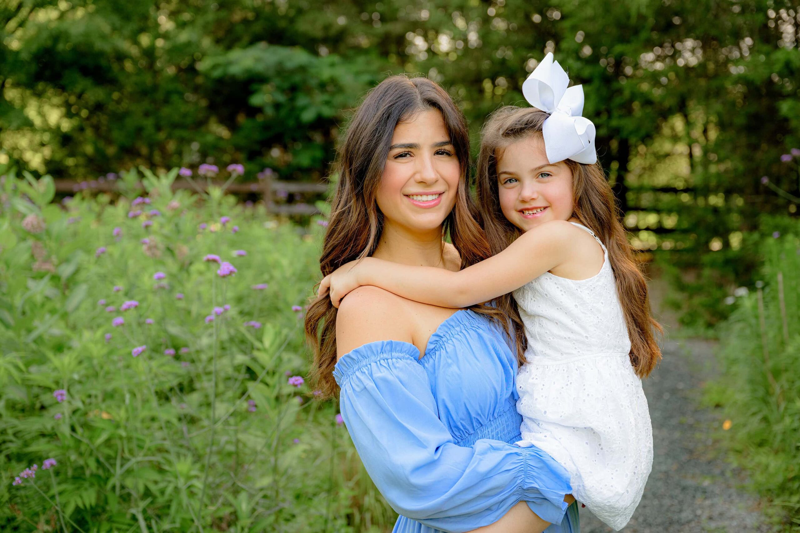 mother wearing a blue dress holds her daughter wearing white with a white bow in a flower garden in waxhaw nc close to pt office