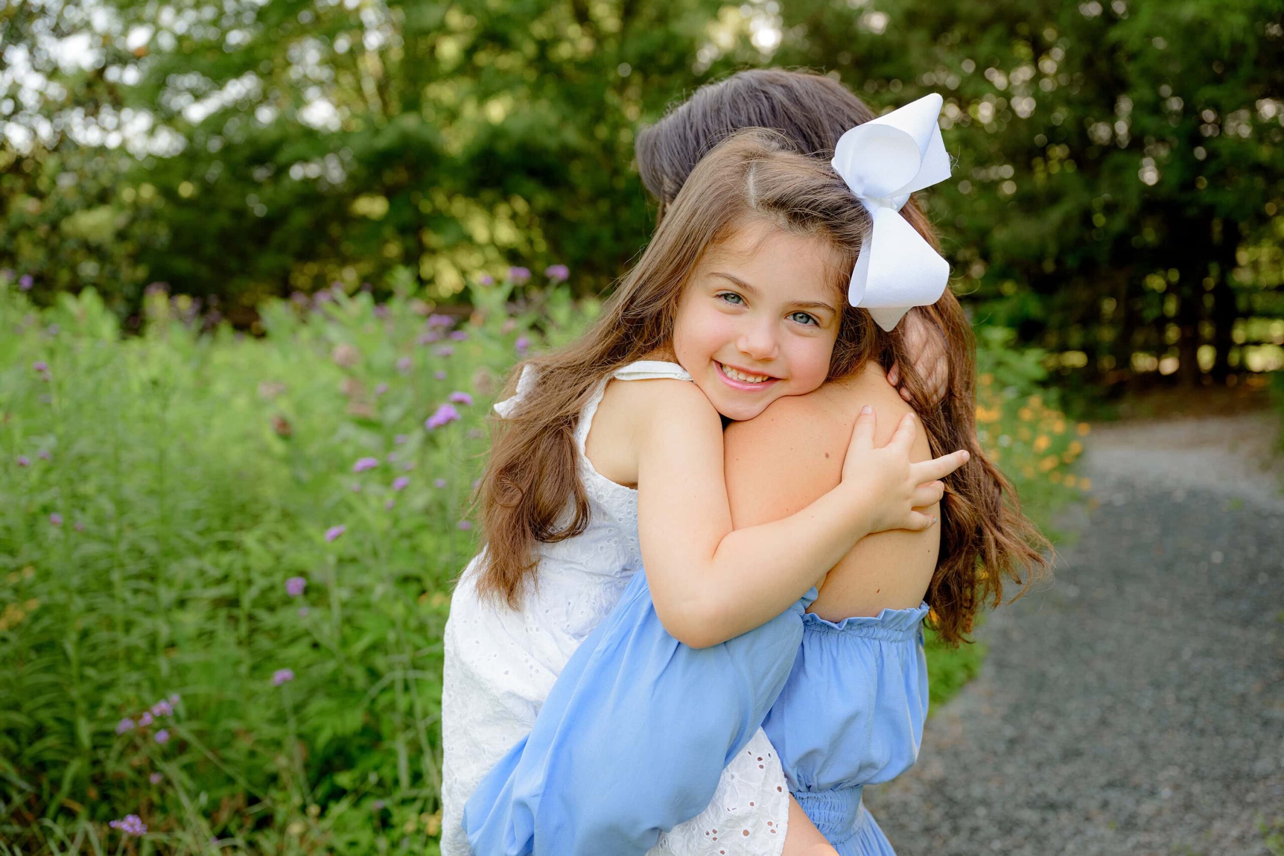 daughter wearing a white dress is being held by her mom smiling with her head on mom's shoulder in a waxhaw area park