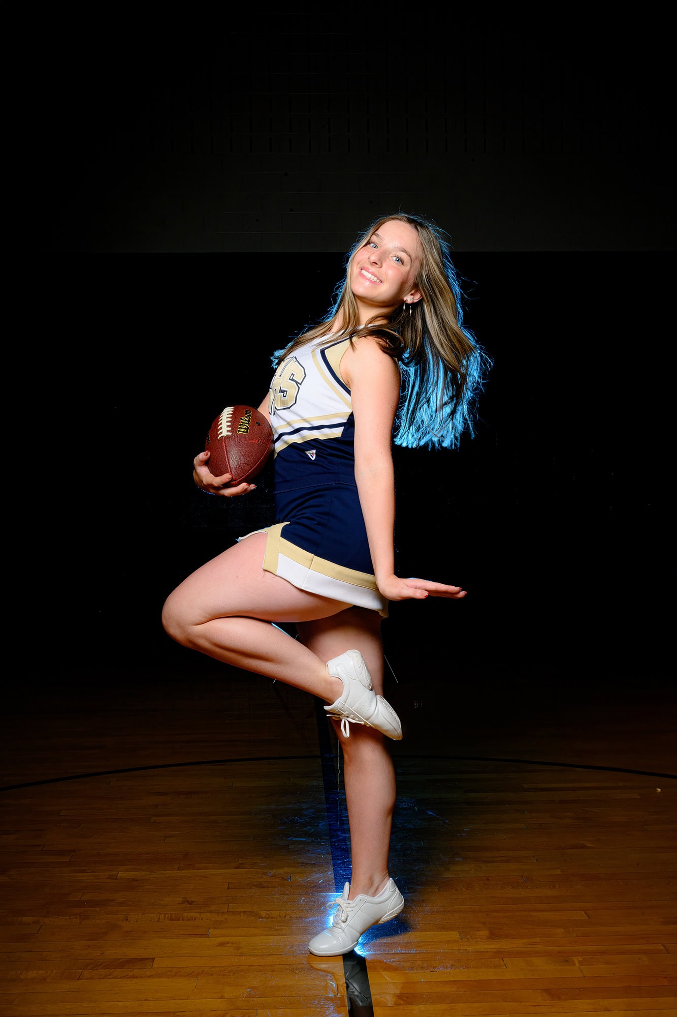 cheerleader posing in her high school gym for media day holding a football