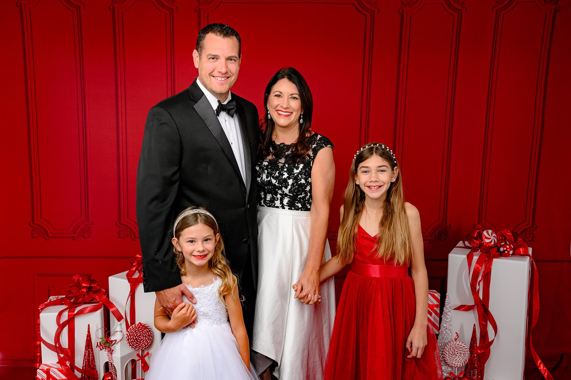family of four dressed up in reds blacks and whites in a red holiday set up with white presents