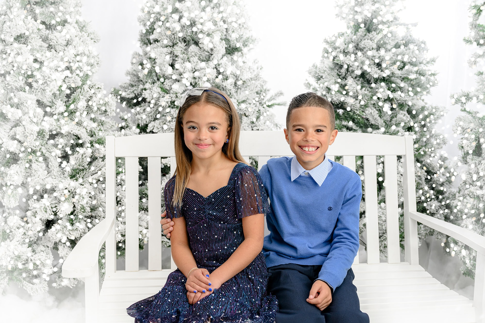 brother and sister in blue sitting on a white bench with holiday trees around them