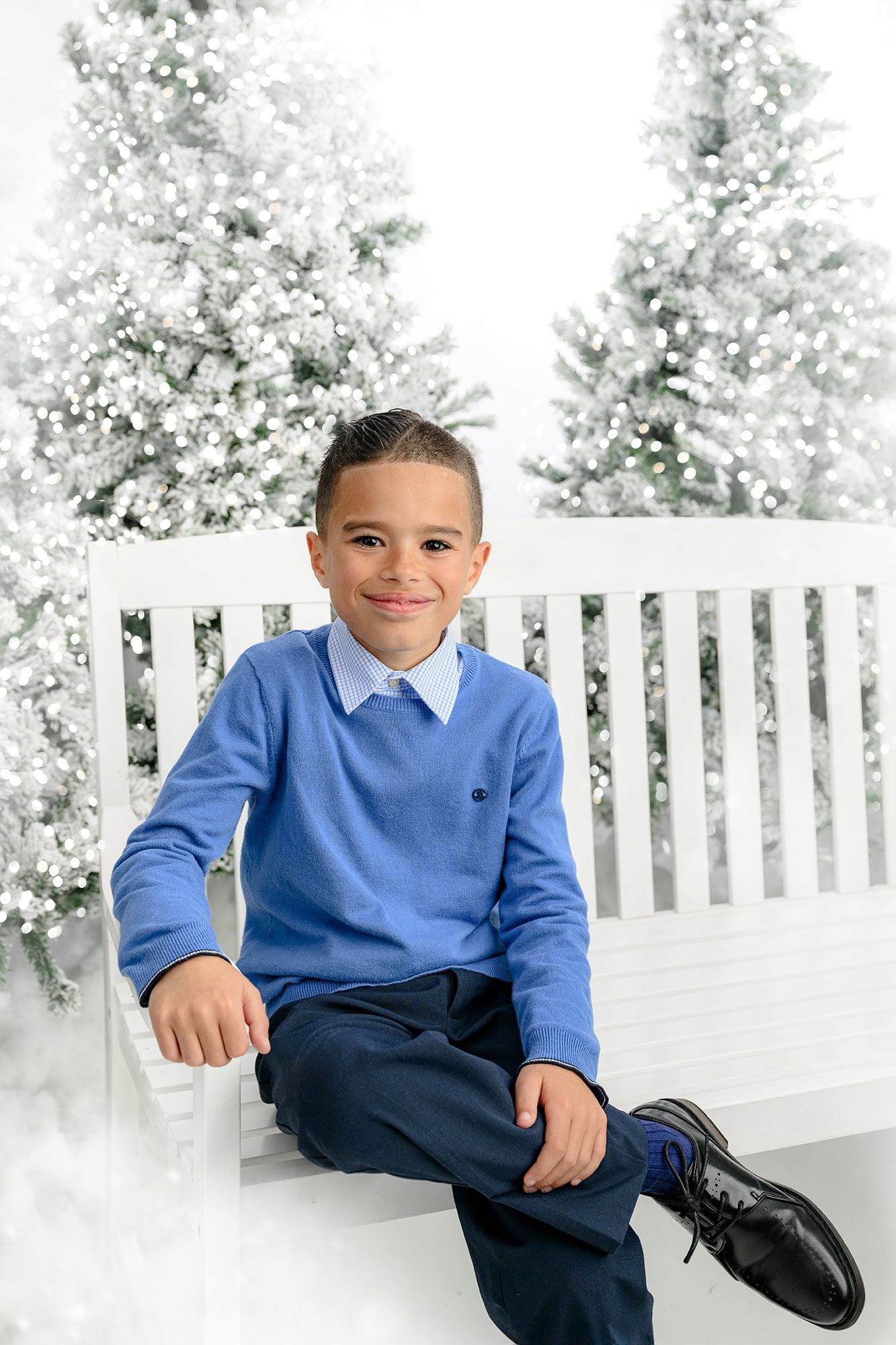 boy dressed in blue sitting on a white bench with flocked christmas trees around him
