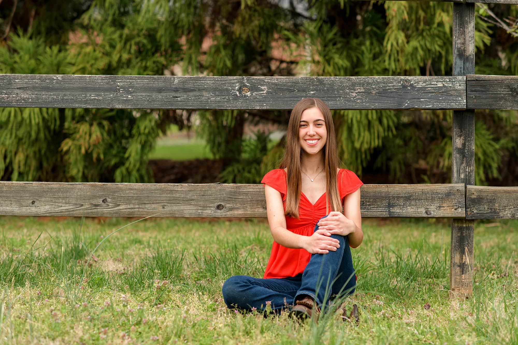 dark hair graduate sits against fenceline wearing a red shirt and jeans and brown boots