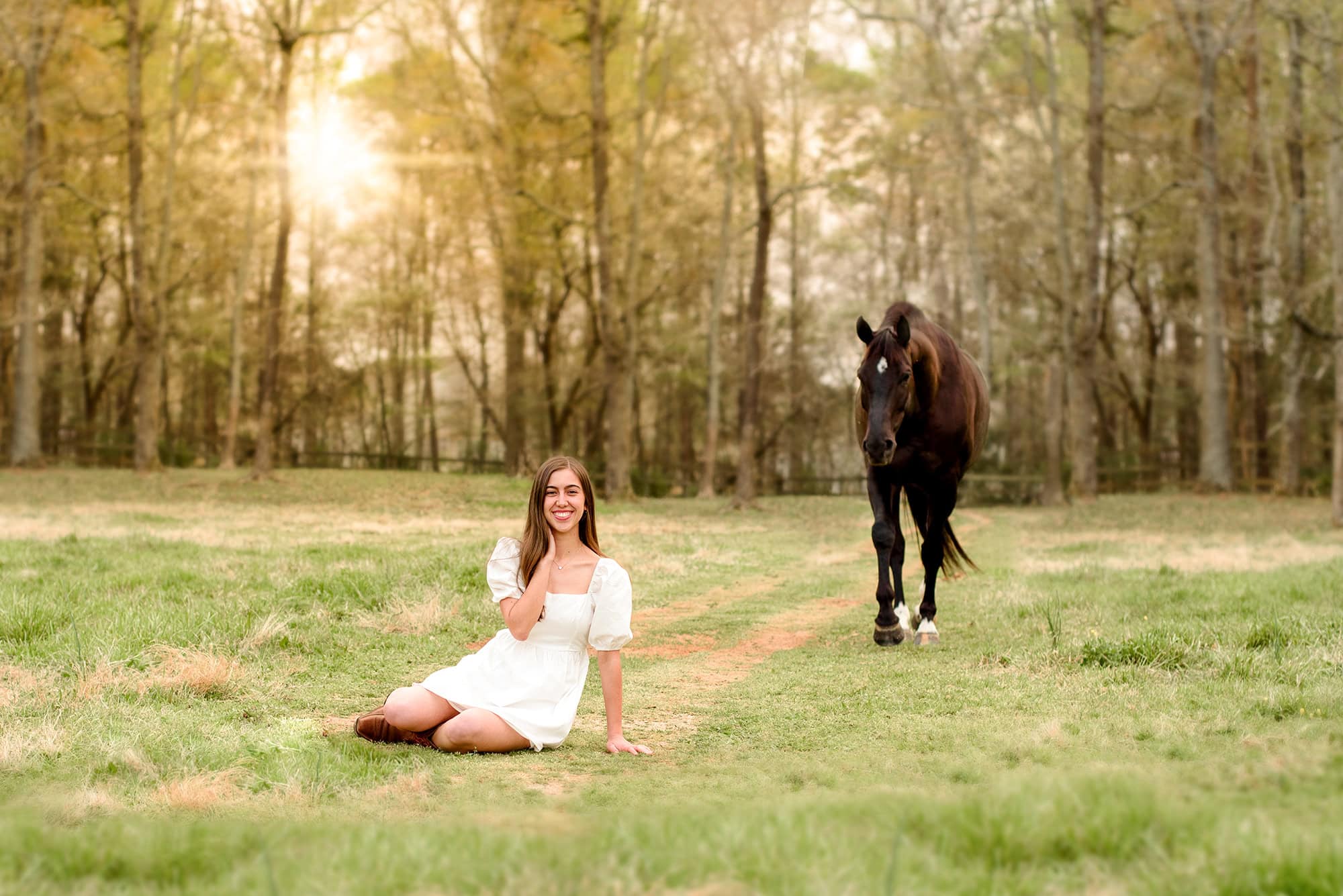 charlotte senior sits in field wearing white dress and boots with her horse behind her and sun setting in a field