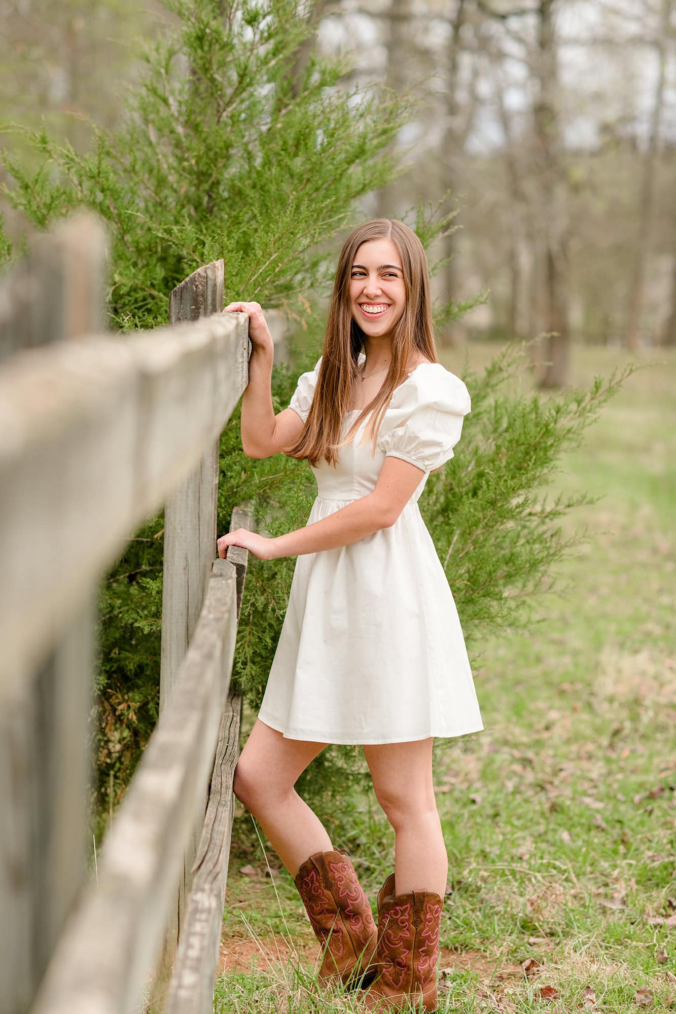 dark hair charlotte senior stands in white dress and brown boots against fenceline