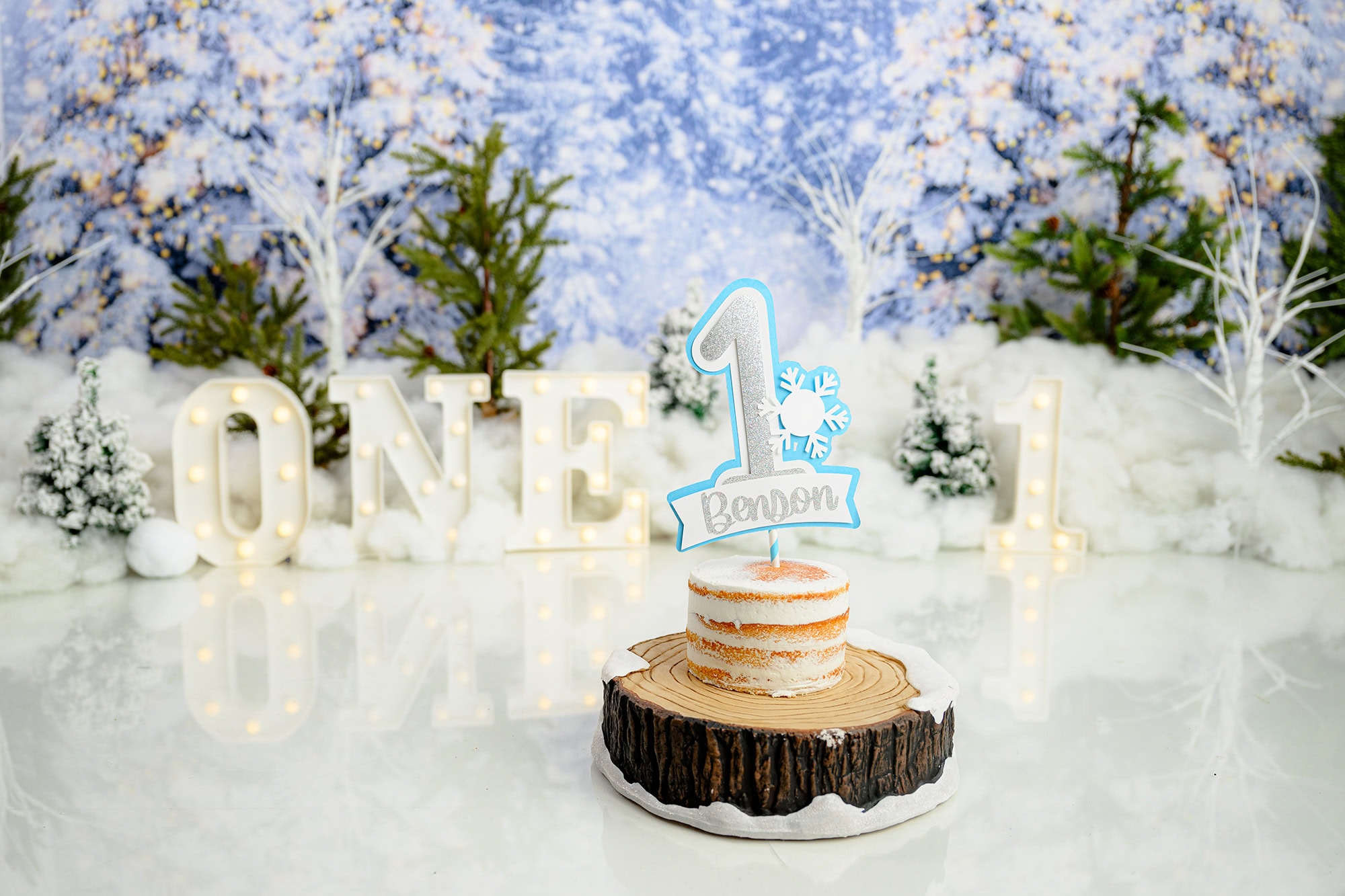 lightly iced cake on a wooden log cake stand with a winter onederland cake smash scene behind 