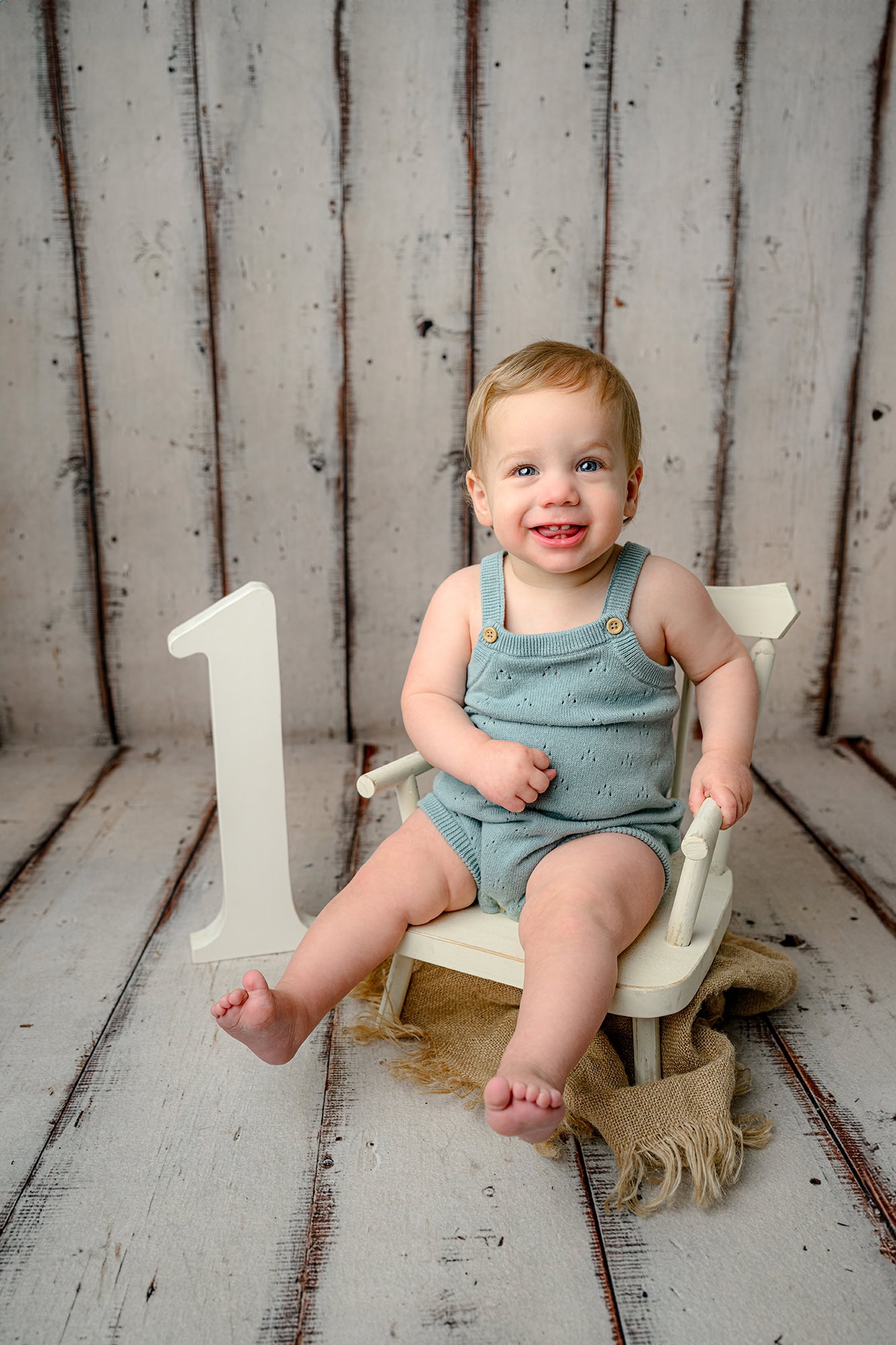 charlotte boy sits in a blue romper in a cream colored chair with a large number one beside him for milestone photos
