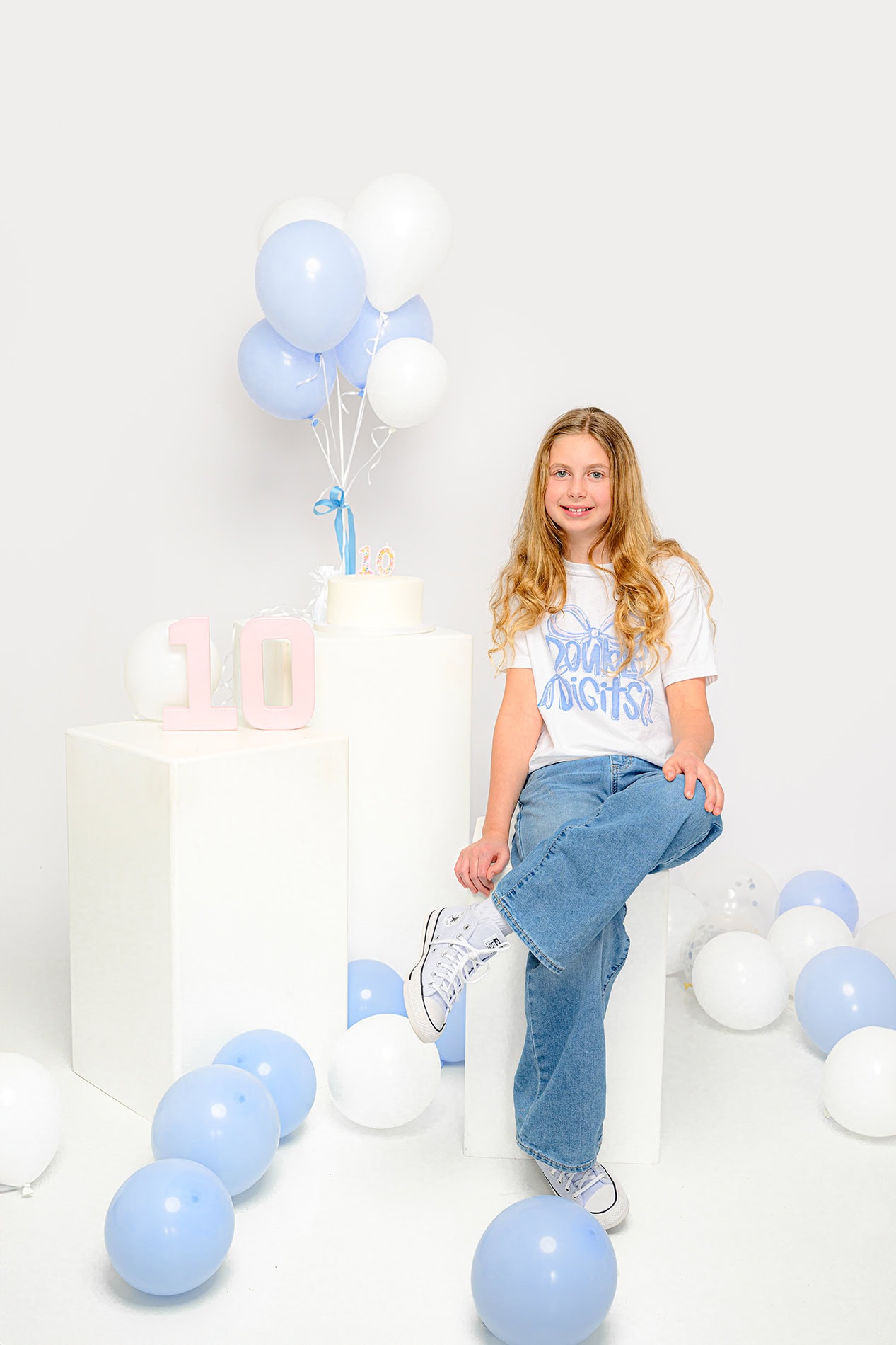 blonde girl sitting on a white stool with birthday decorations and balloons around her