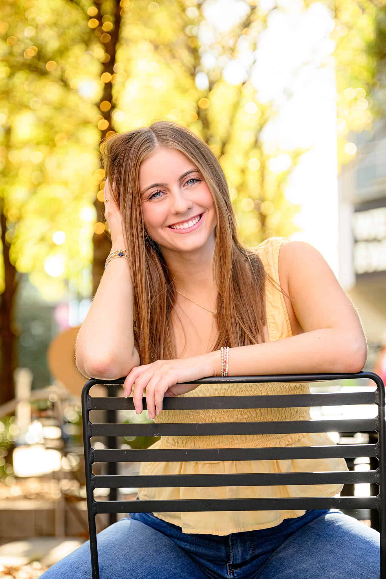 uptown graduate session photo of senior wearing a yellow top sitting in chair with yellow trees behind her