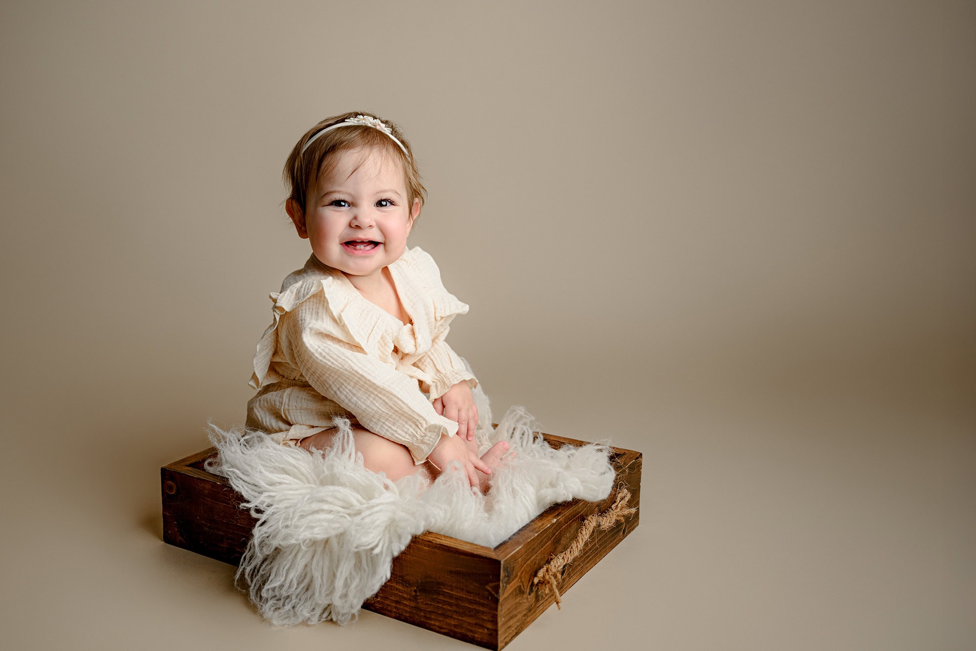 one year old girl sits in wooden box wearing a cream romper