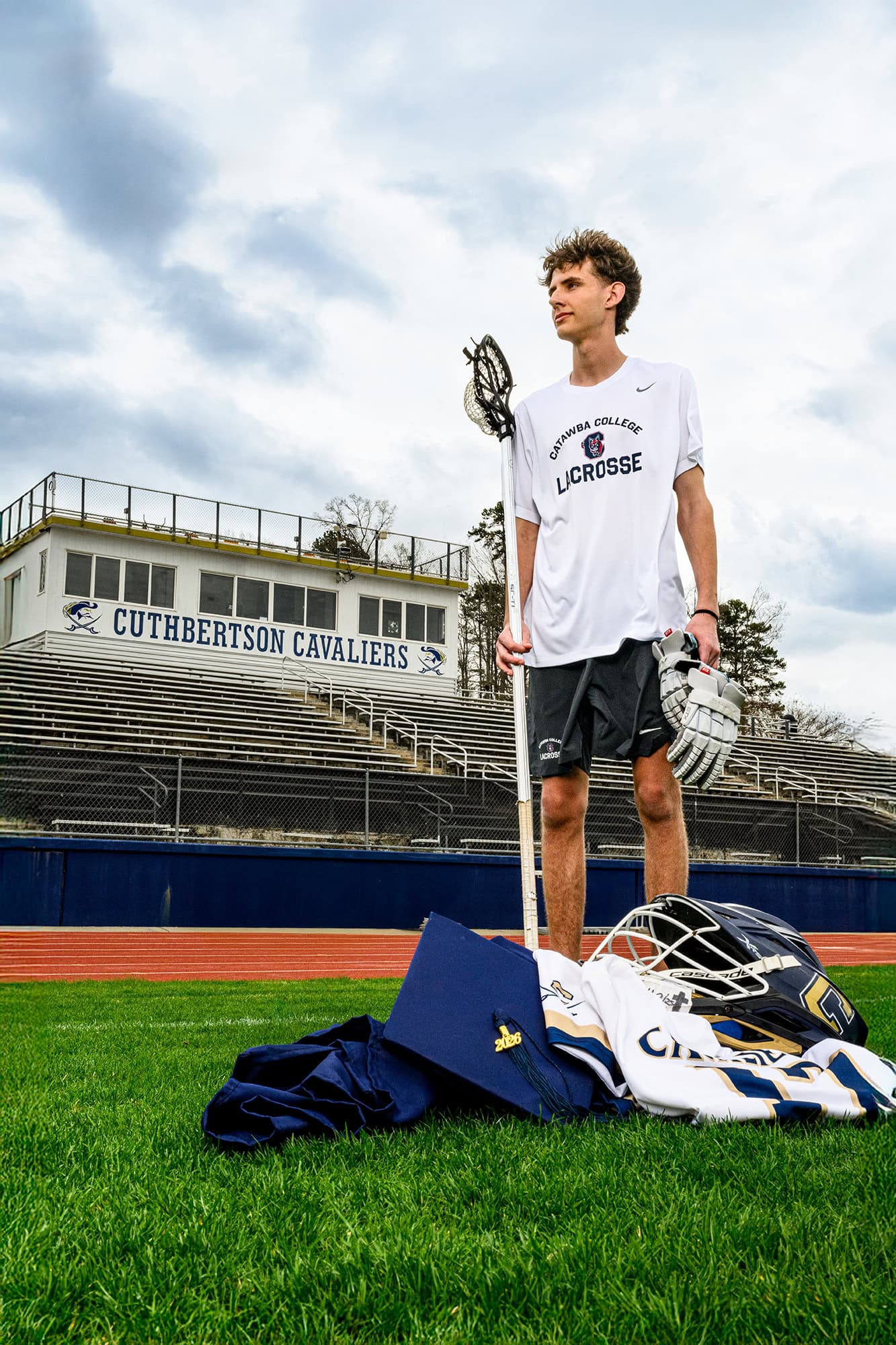 nod to future plans for this charlotte lacrosse player wearing his college gear with his high school gear and cap and gown on the ground in front of him