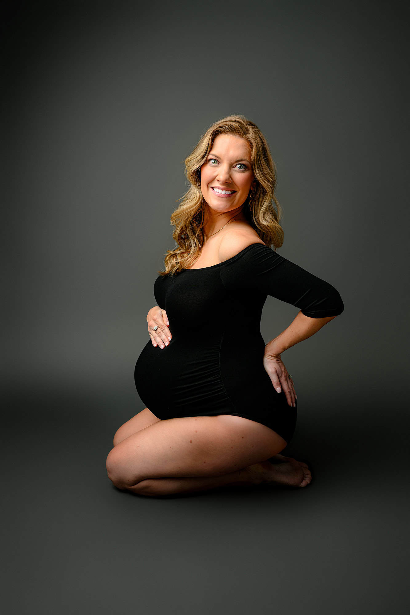 blonde mom to be in a black bodysuit sitting on her knees with her hand on her belly and hip