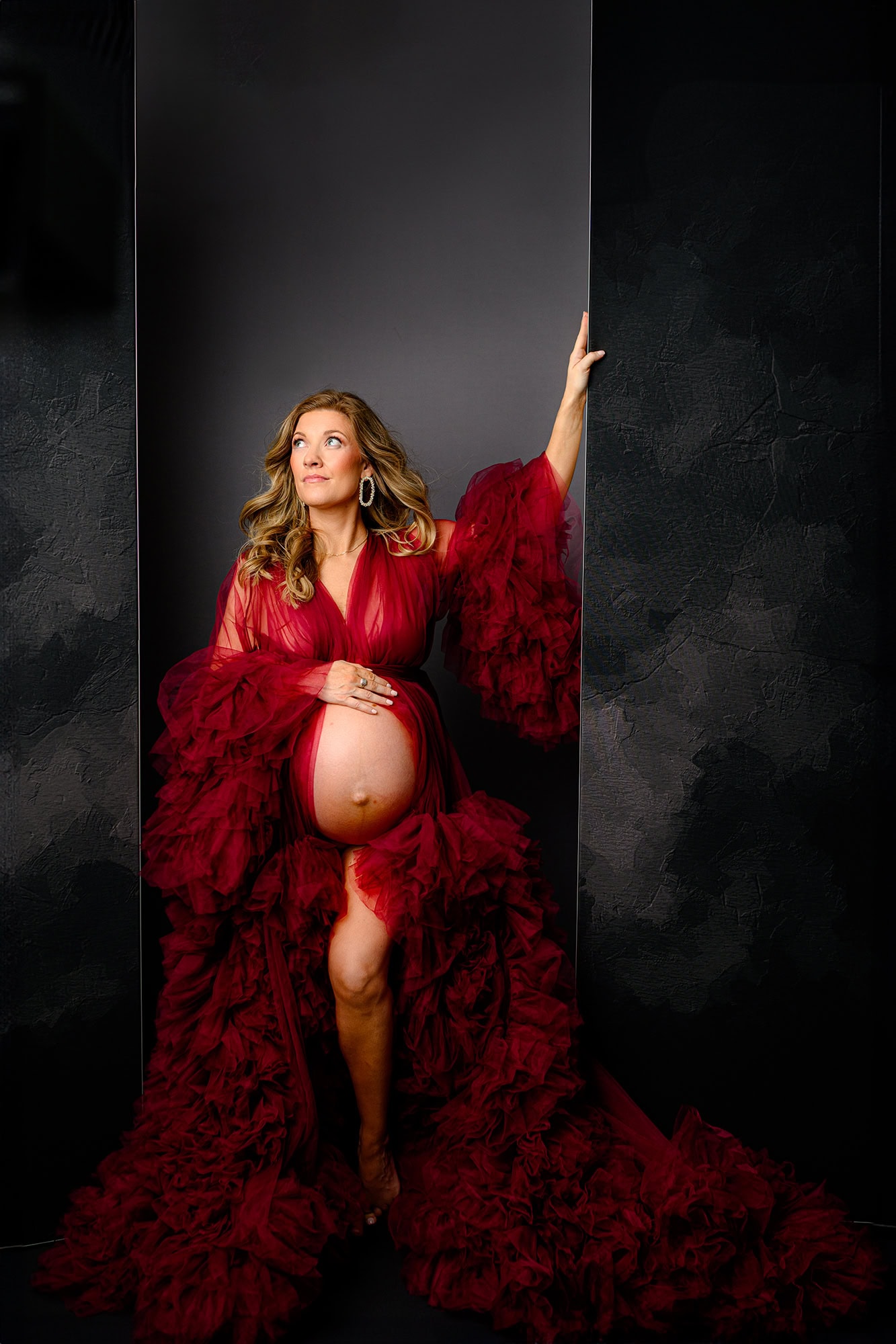 mom to be in a dramatic red dress posing with belly out and glam hair and makeup for her studio maternity session
