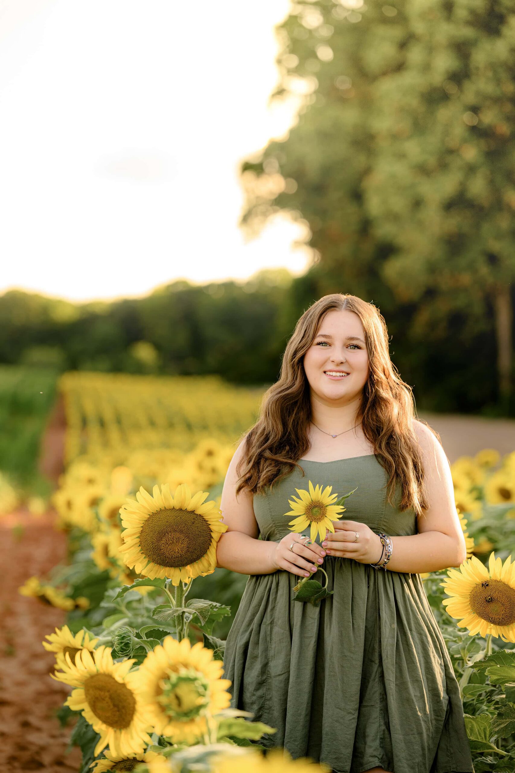 dark hair grad standing in a green dress holding a sunflower in a large field