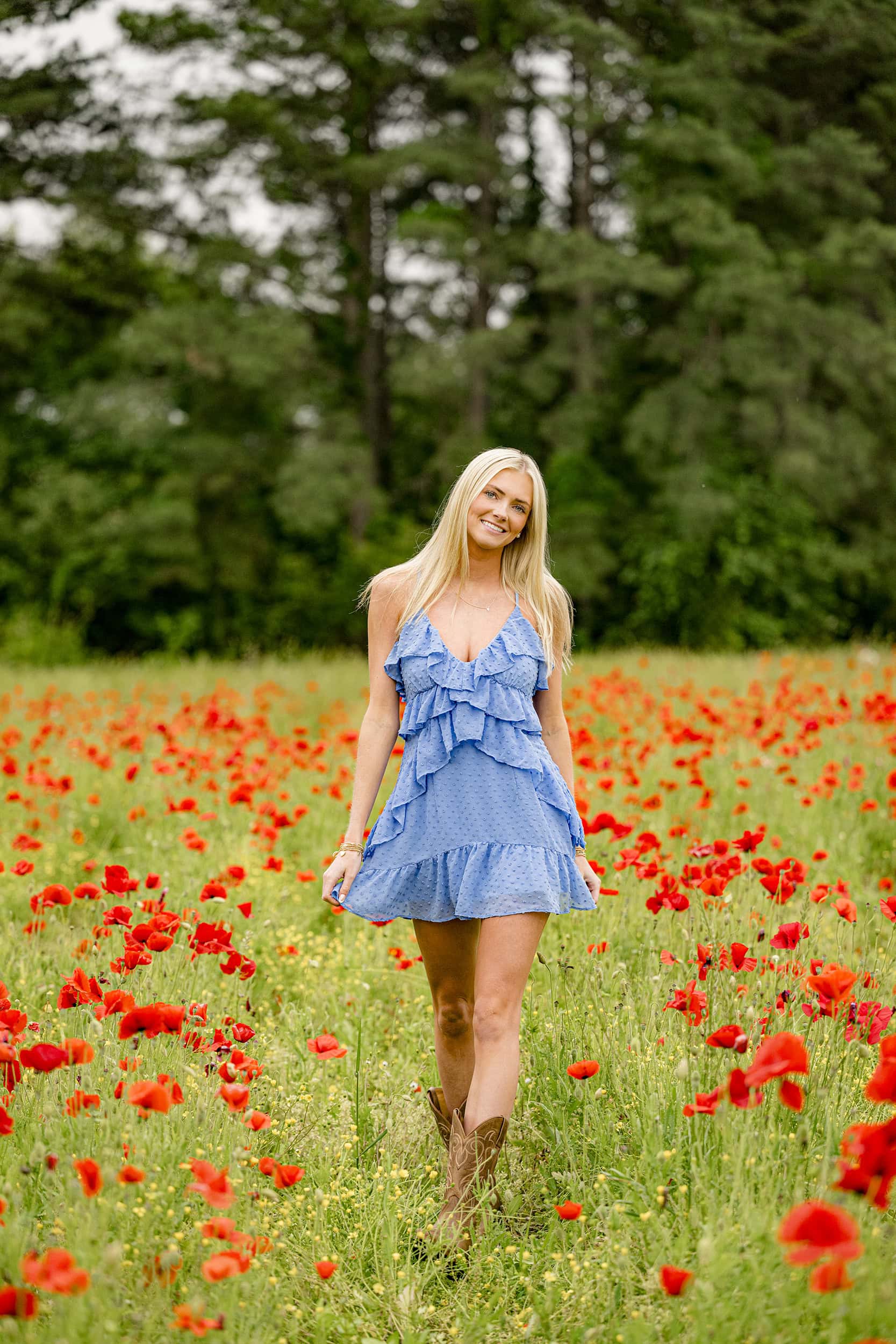 blonde charlotte senior holds her blue dress standing in a red poppy field