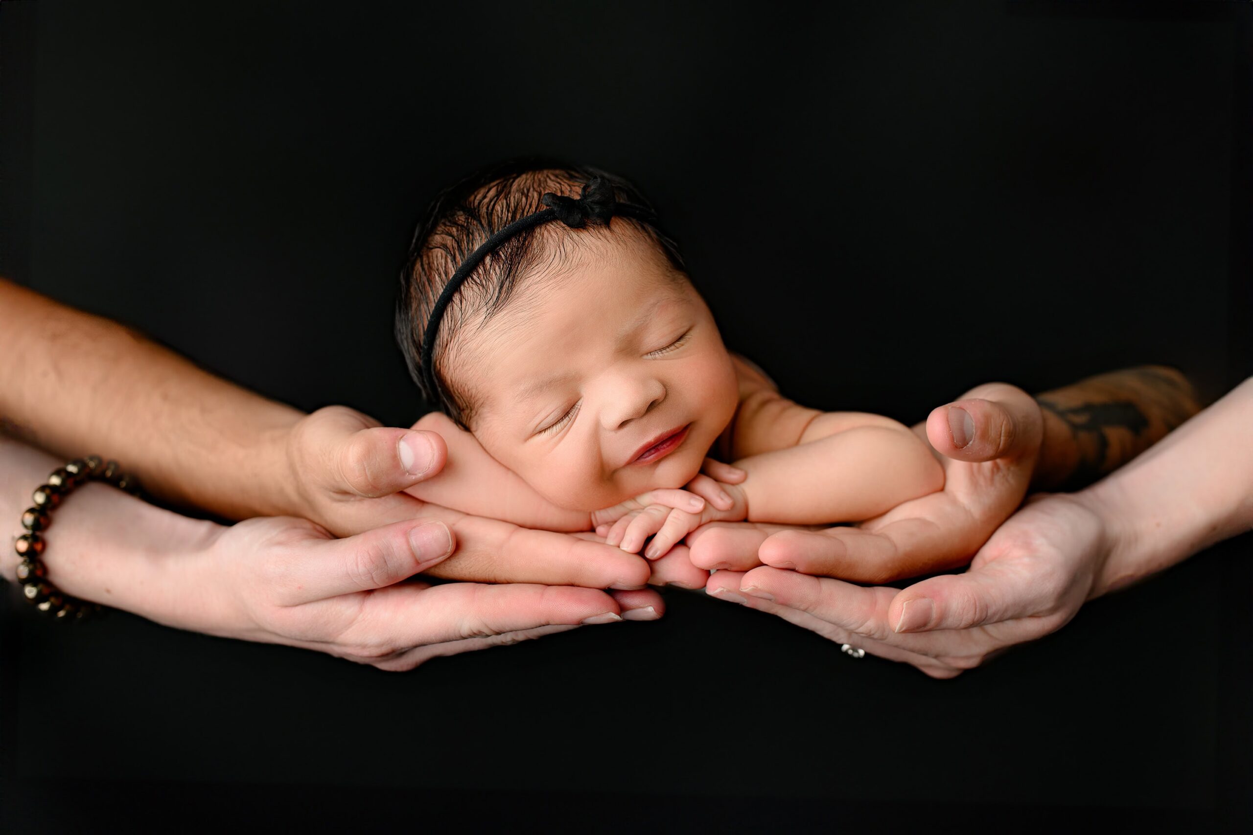pose of newborn girl laying in her parents hands on a dramatic blackbackground