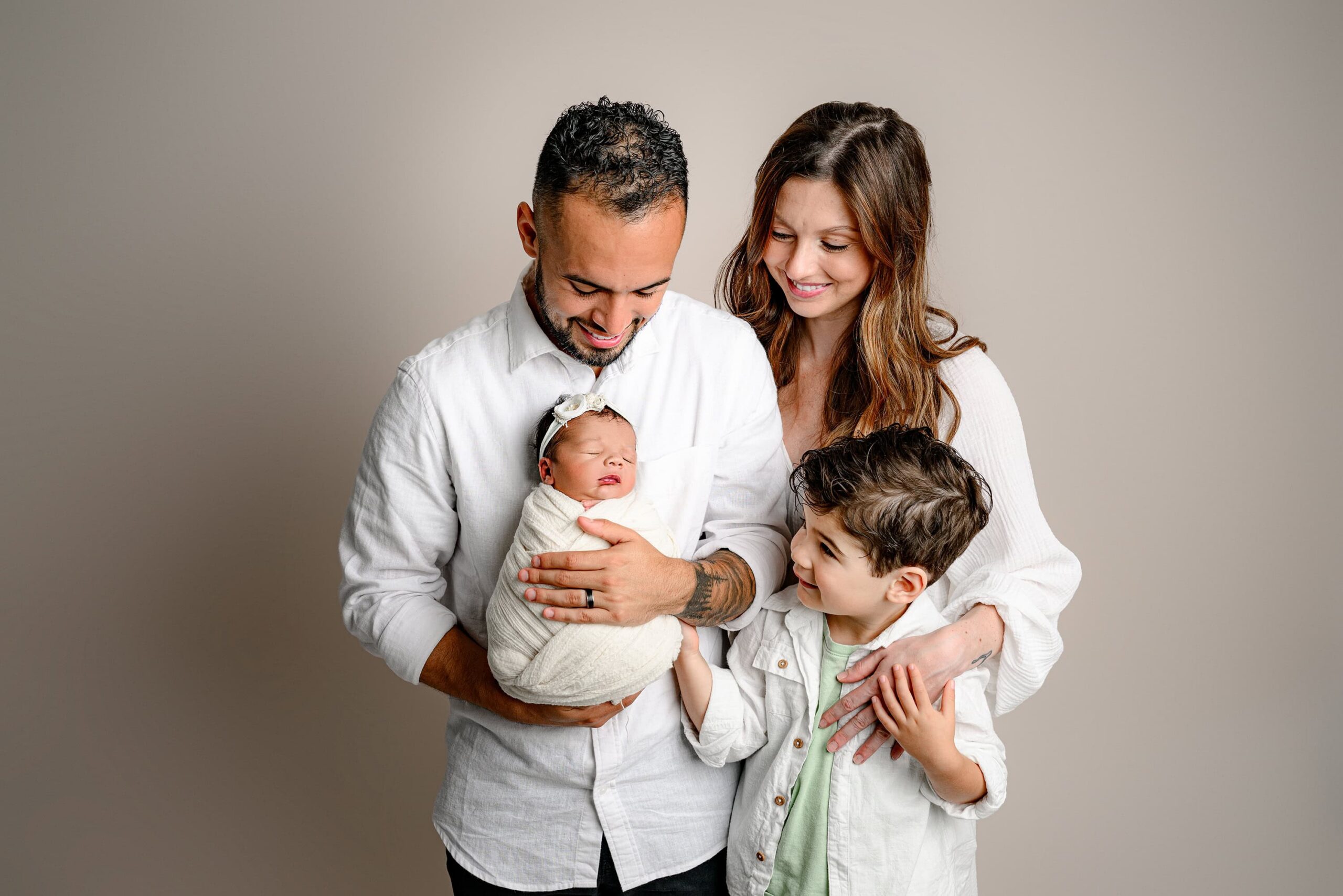 Family of four dressed in neutral colors all looking at their swaddled newborn daughter