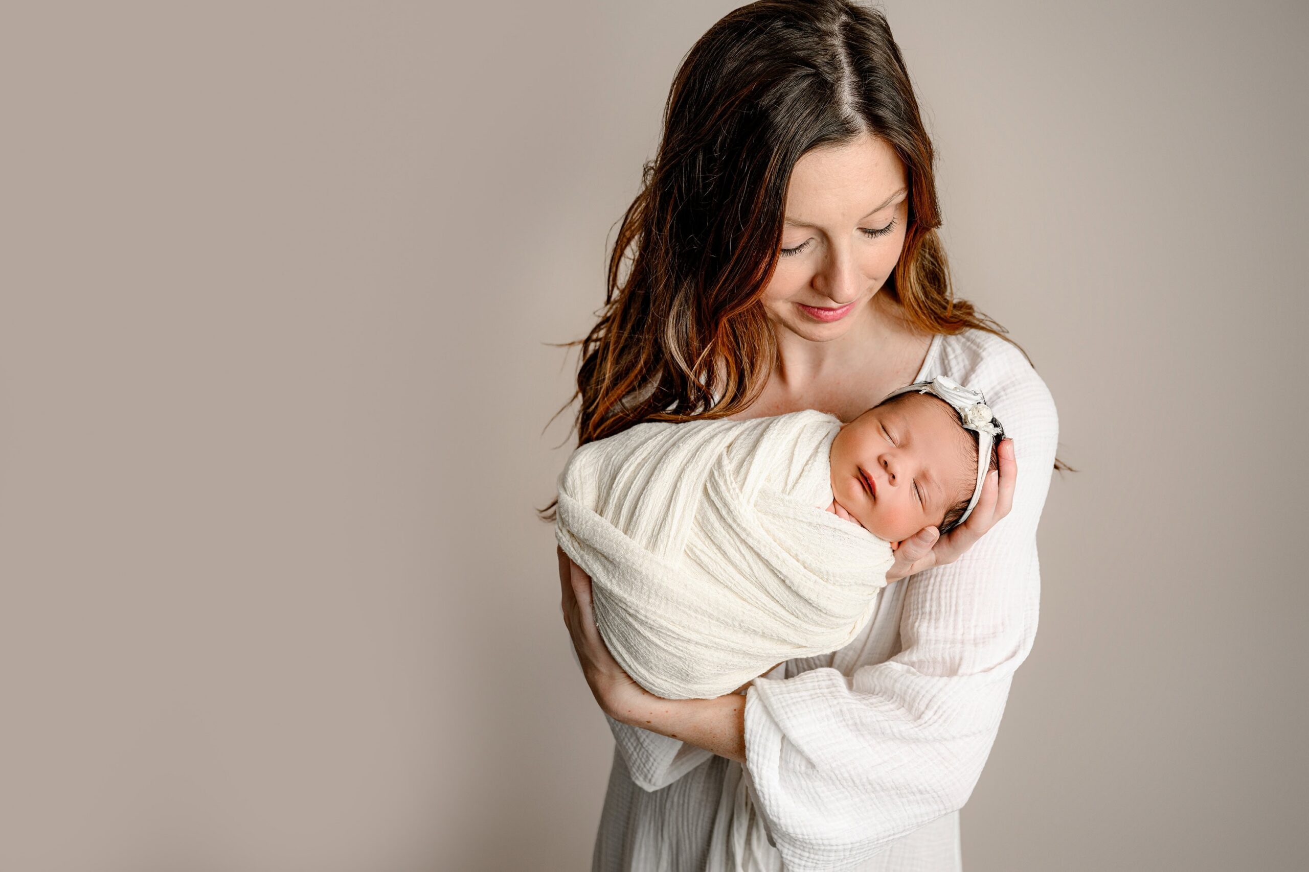 charlotte mother wearing a white dress cradles her newborn daughter wrapped in cream during their newborn family photos