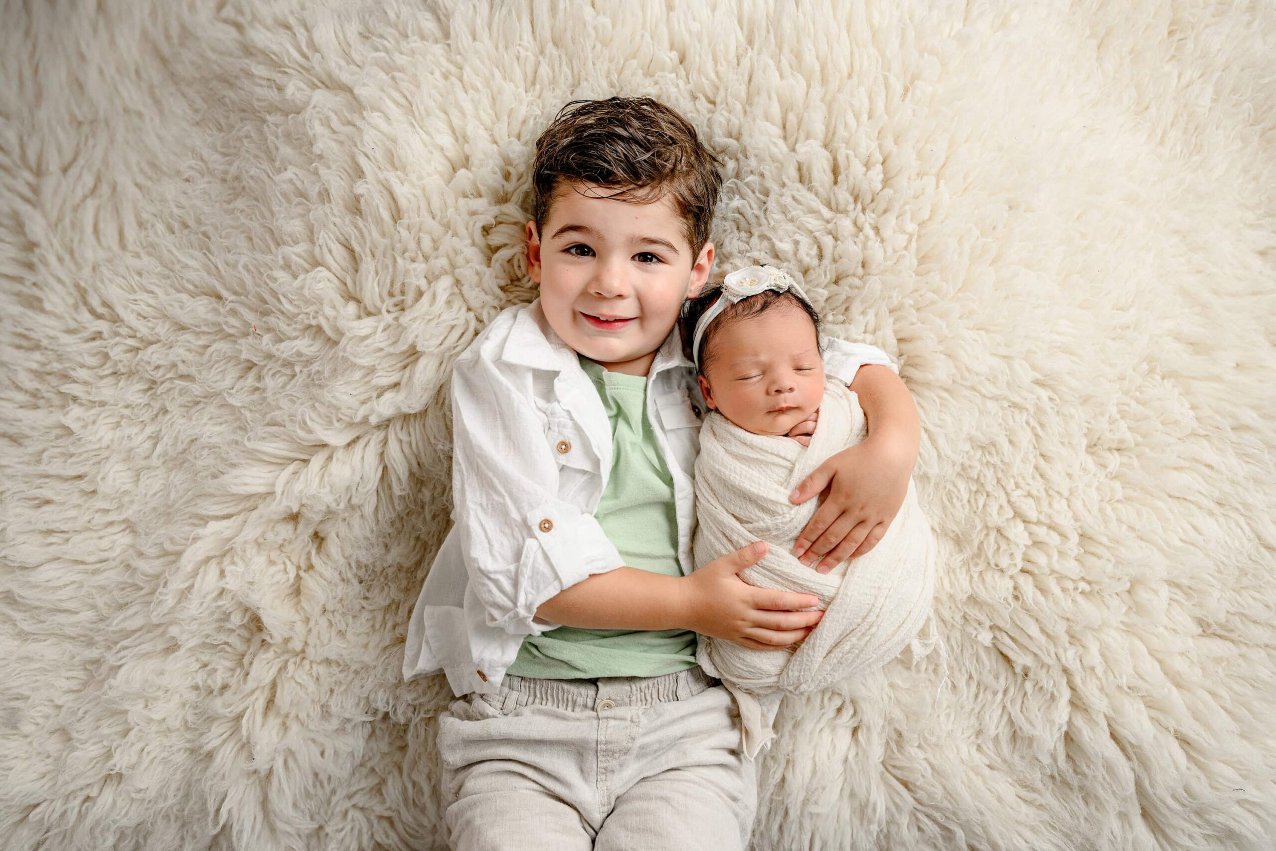 little boy wearing a green shirt with a white shirt over it cuddling his newborn sister during family photos