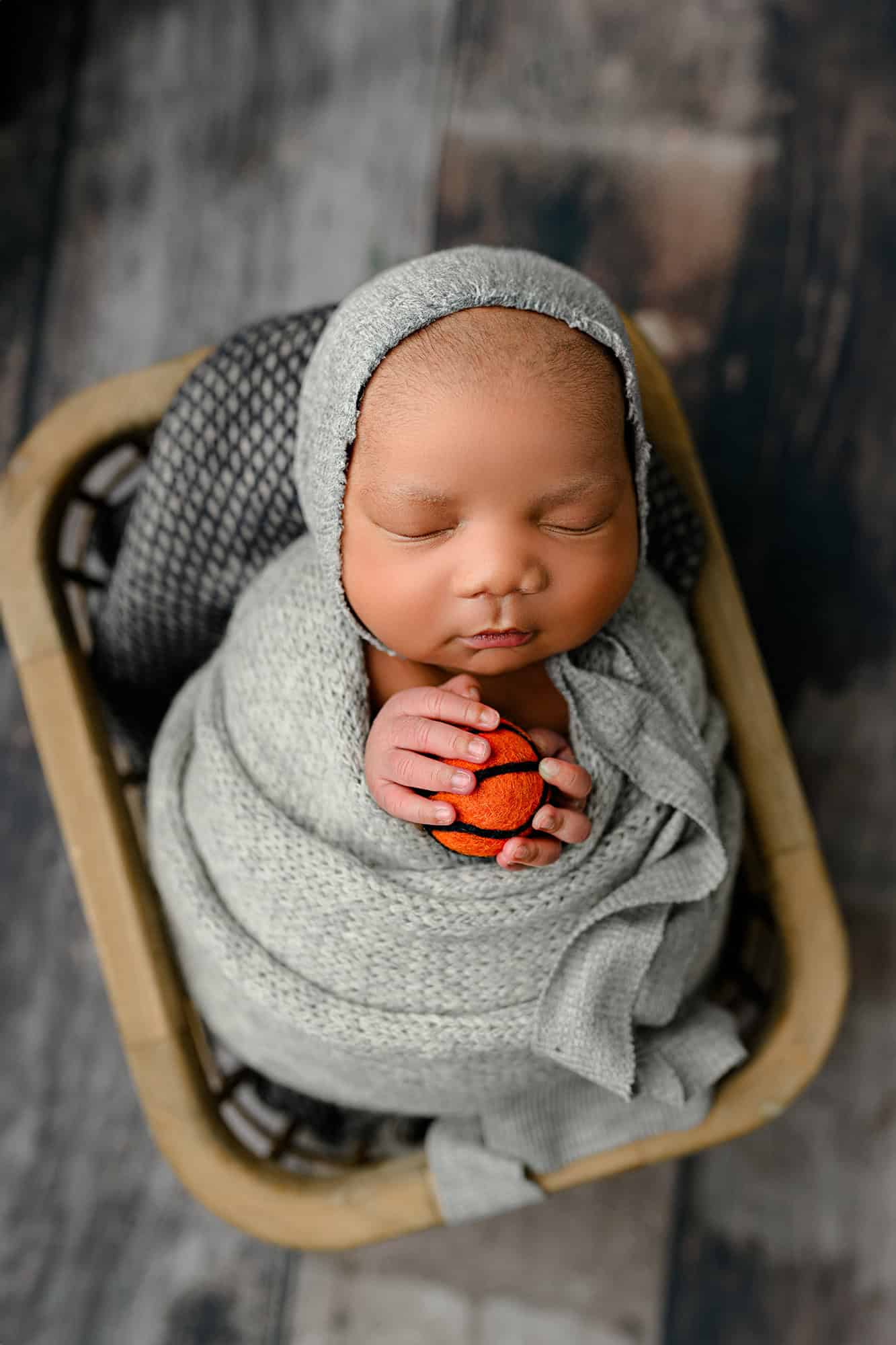newborn baby boy wrapped in a grey wrap holding a basketball