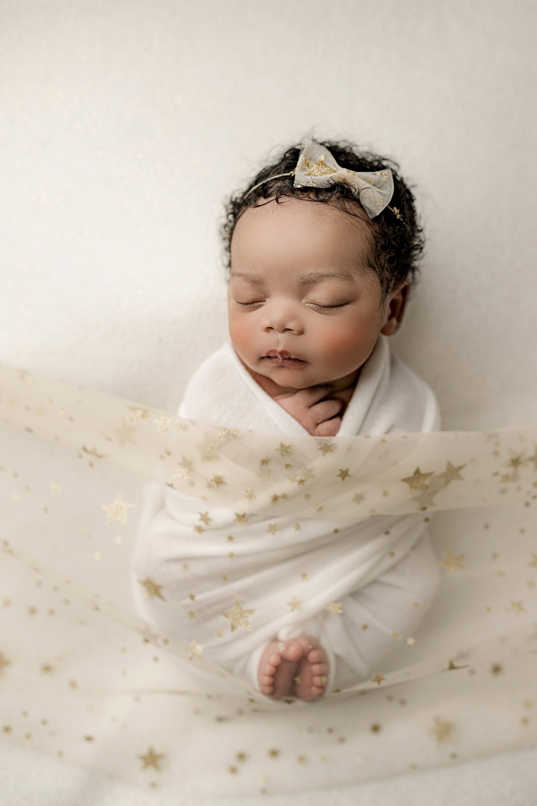 newborn baby girl 5 weeks old wrapped in a white wrap with her toes peeking out with a gold start cover and bow