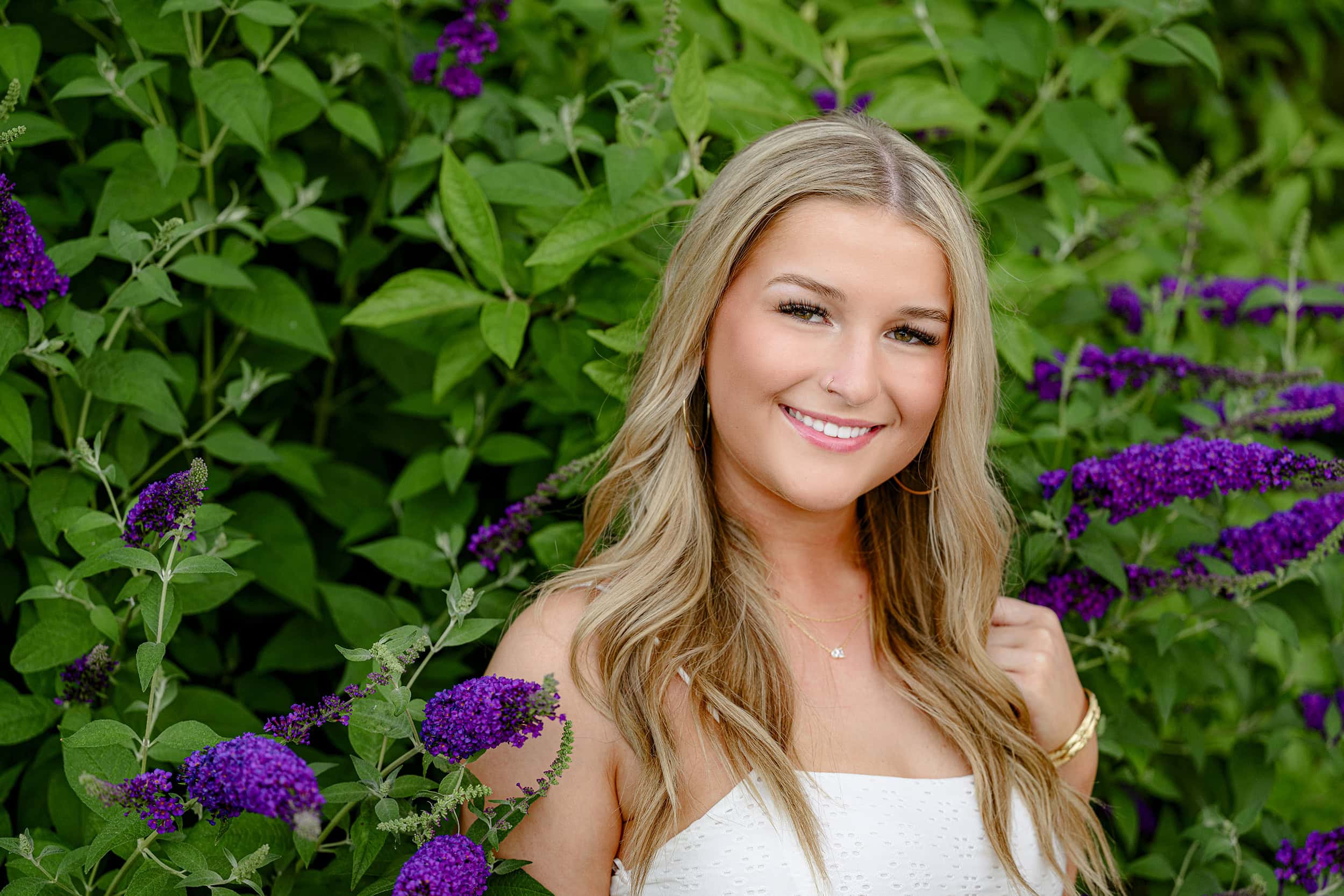 blond senior in high school wearing a white dress amongst purple blooms with her eyelashes and eyes beautifully highlighted with natural makeup