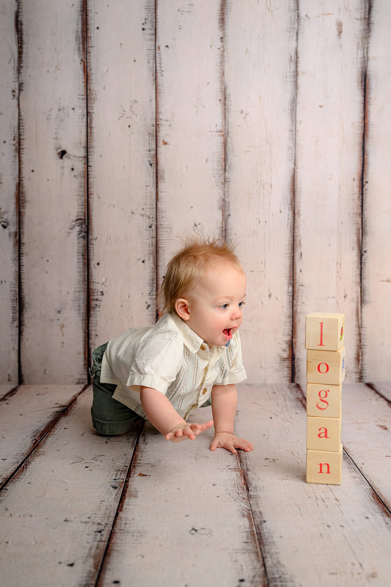 one year old boy crawling toward a stack of wooden blocks wearing a cream shirt and green shorts
