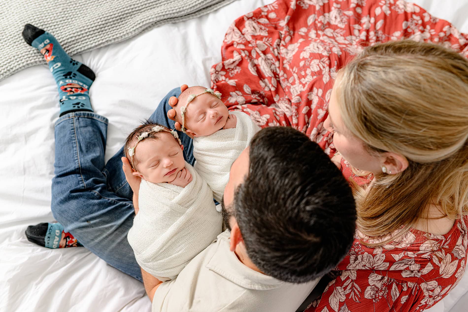 two parents sitting on their bed holding both of their twin newborn daughters