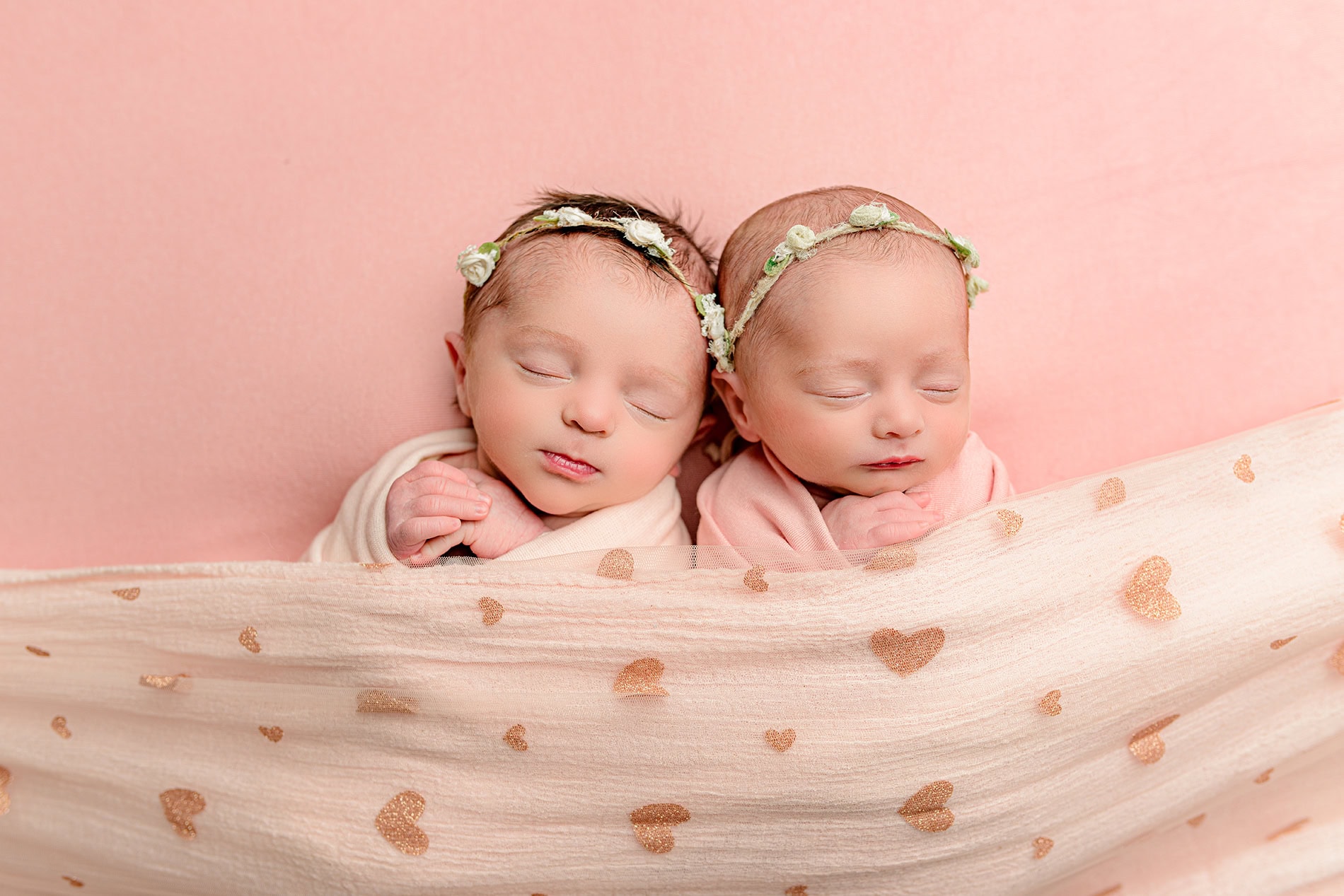 newborn twin girls laying side by side wrapped in pink with floral headbands on during their lifestyle newborn session 