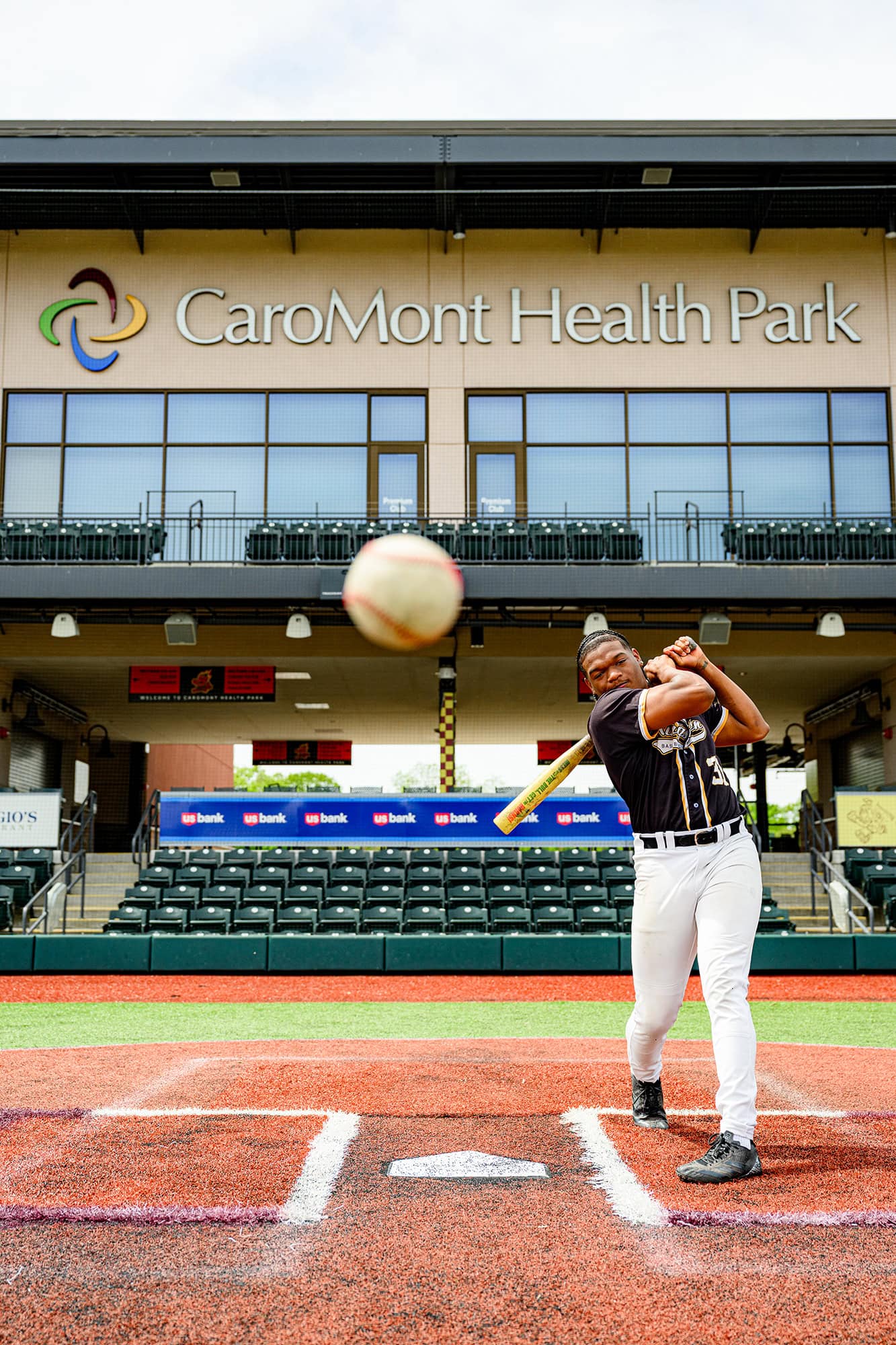 charlotte senior photo of kingsmen academy athlete hitting a baseball with caromont health park logo and stands behind him