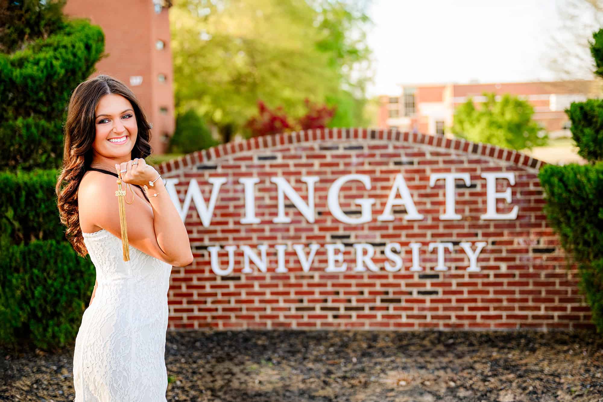 Wingate University graduate stands in front of campus sign holding her graduation tassel and wearing a white dress