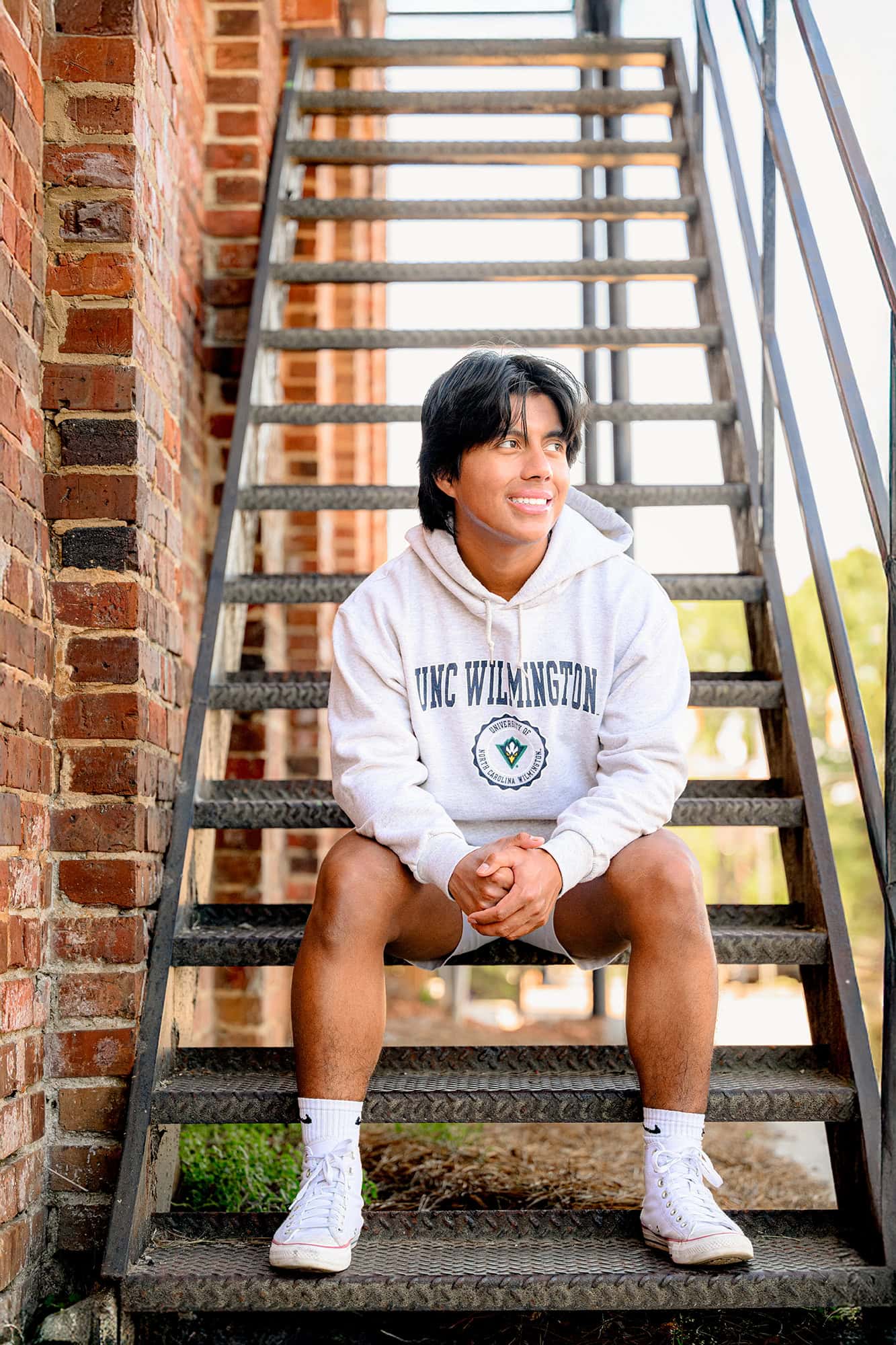 senior boy wearing his college gear leans on metal stairwell in downtown waxhaw, nc in the morning