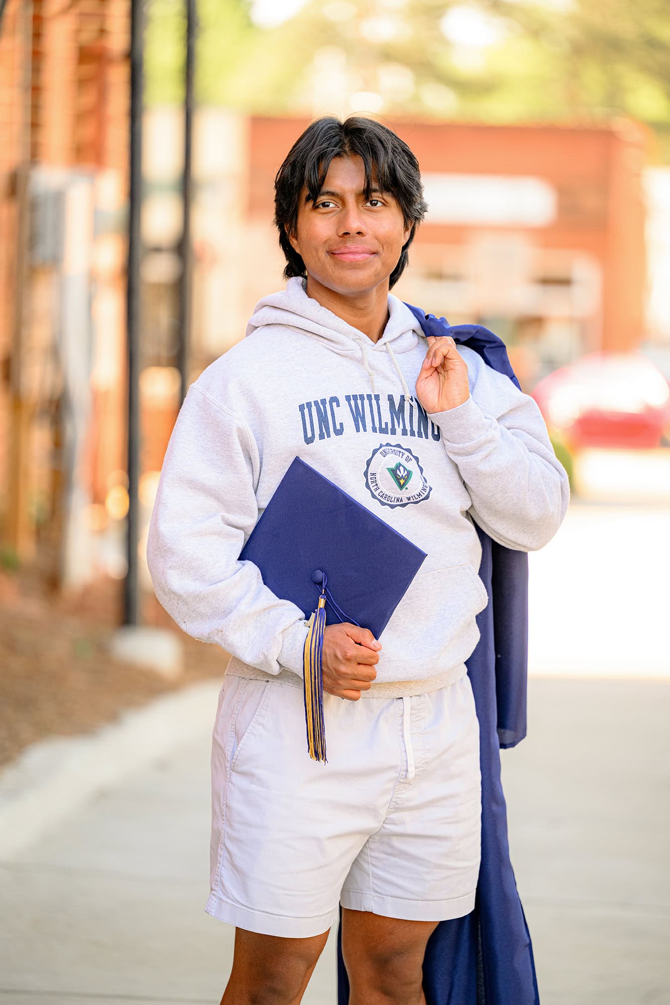 dark hair senior boy wearing a college shirt with his gown over his shoulder holding a graduation cap in downtown waxhaw