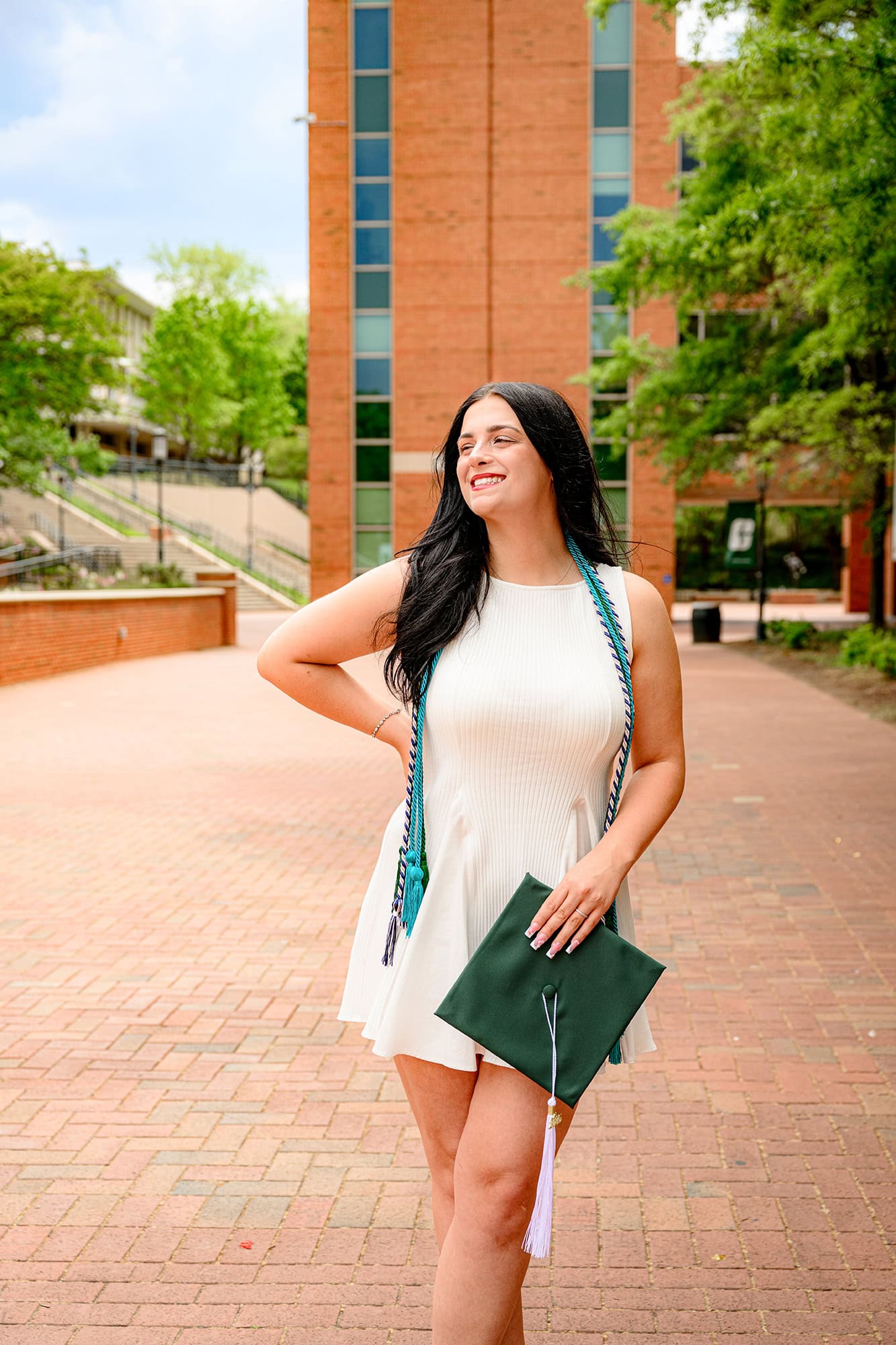 UNC Charlotte grad stands in front of clock towner with her hat in her hand wearing her cords