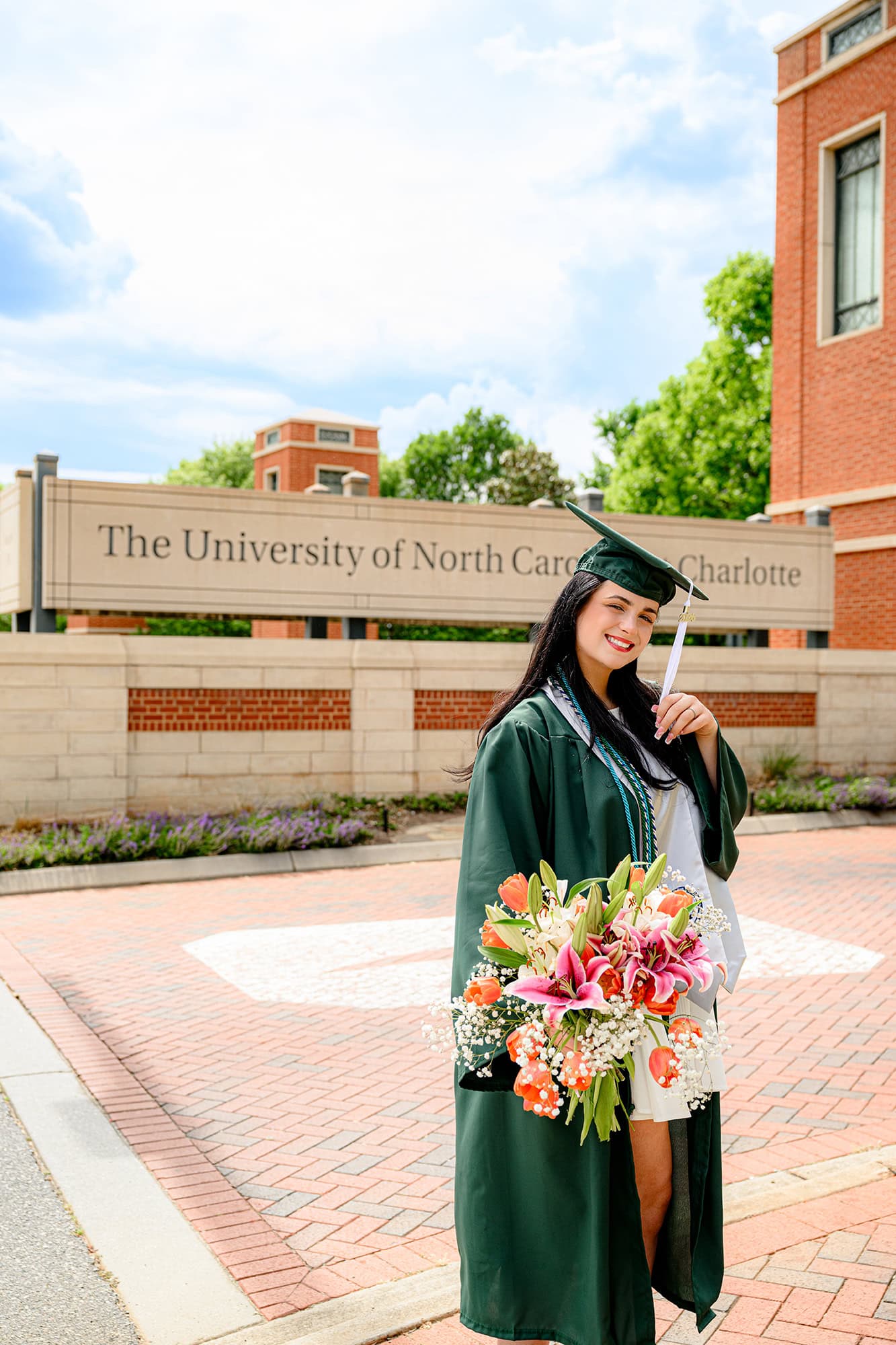 UNC Charlotte grad holds a floral bouquet wearing her full graduation attire and holding her cap tassel in front of UNC sign