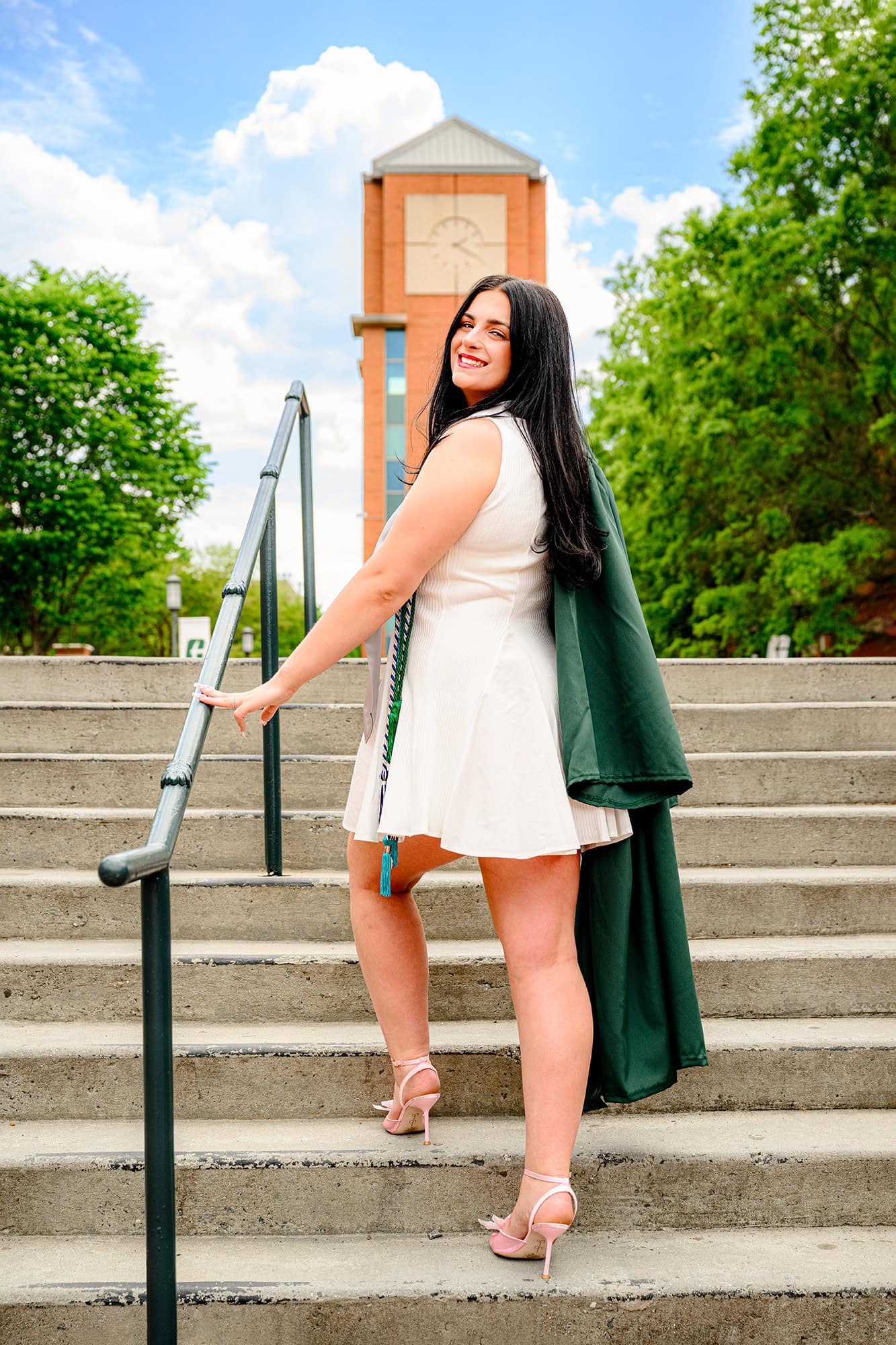 UNC Charlotte senior holding her graduation gown in front of clock tower walking up steps and looking back over her shoulder