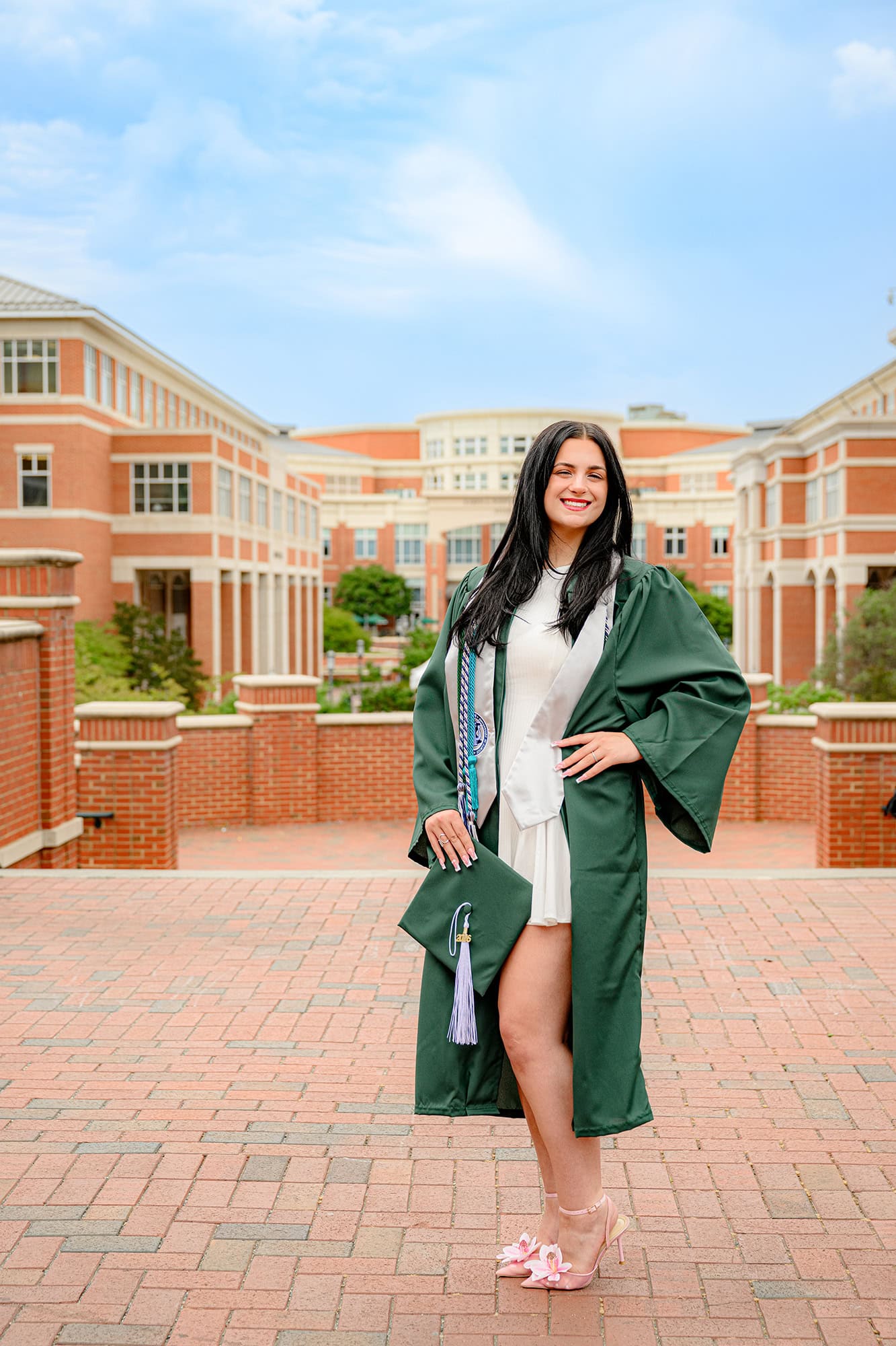 UNC Charlotte senior wearing her graduation gown holding her cap with her hand on her hip in front of student union