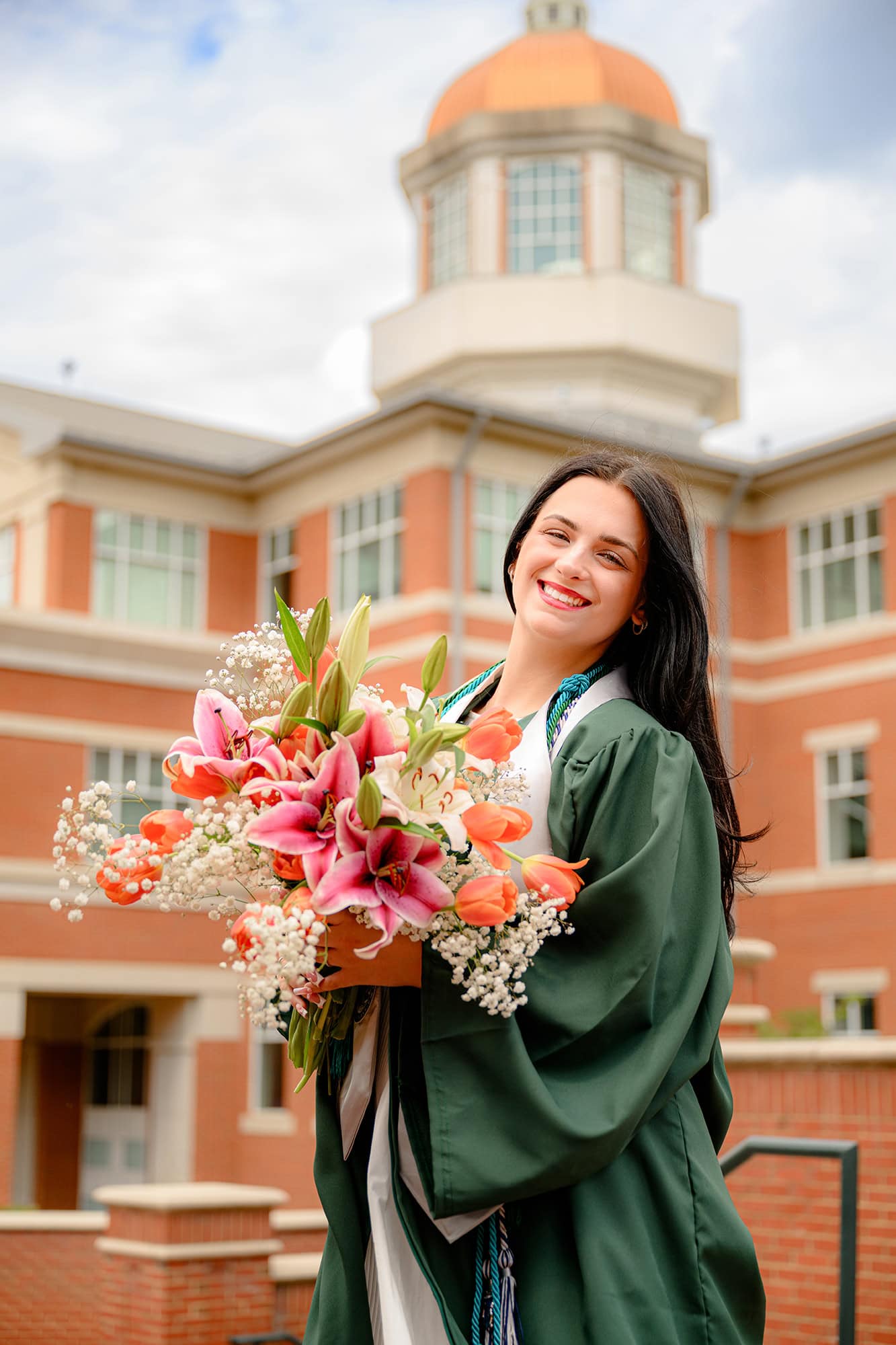 UNC Charlotte grad stands in front of building wearing her full grad gown and cords holding a floral bouquet
