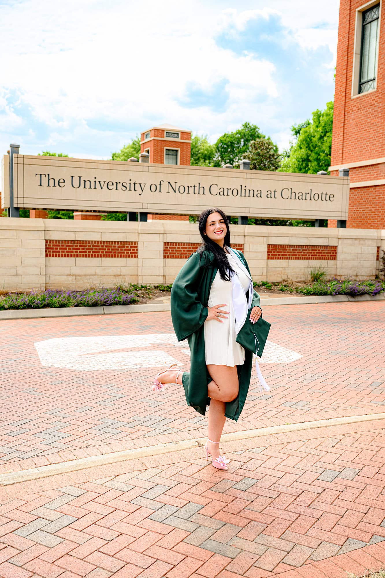 Dark hair UNC Charlotte grad standing in front of the UNC campus sign wearing her grad gown and holding her cap and tassel.