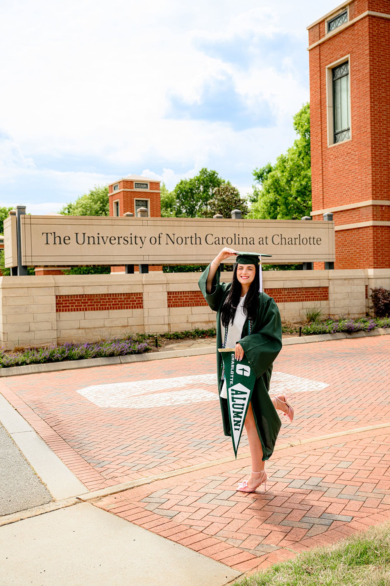Dark hair UNC Charlotte grad wearing her full graduation attire and holding the tip of her cap and an alumni pennant in front of the campus sign
