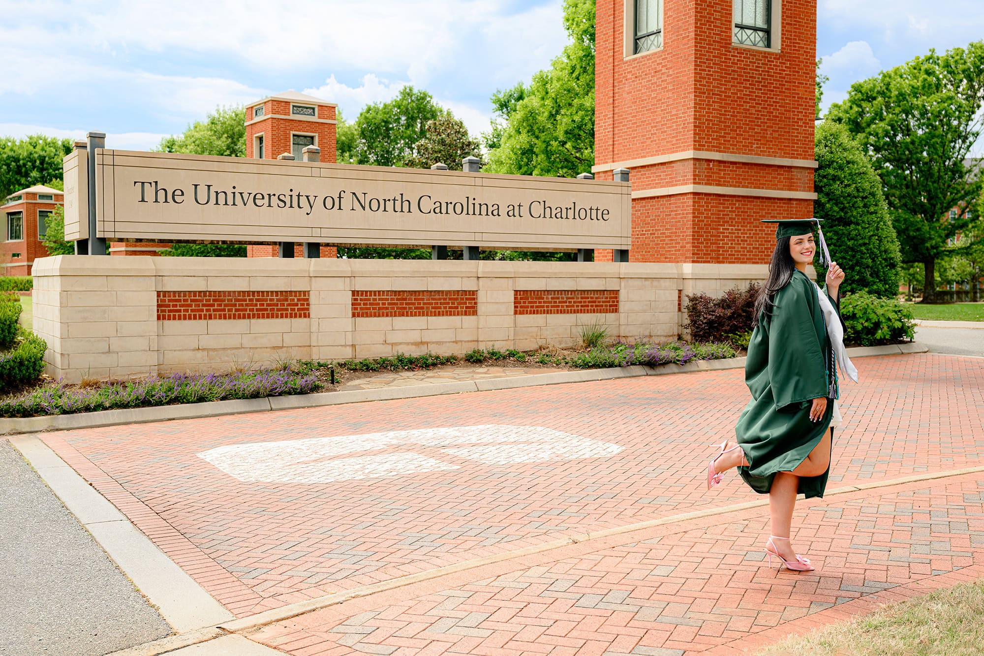 Dark hair UNC Charlotte grad posing with her foot up in front of UNC Charlotte campus sign wearing her full graduation attire.