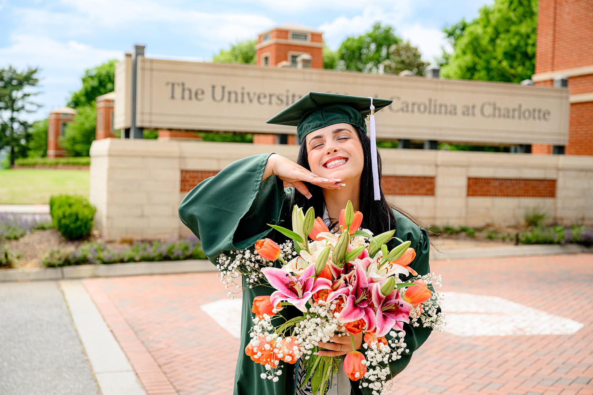 Dark hair UNC Charlotte grad wearing her full graduation attire posing with her hand under her chin celebrating her graduation