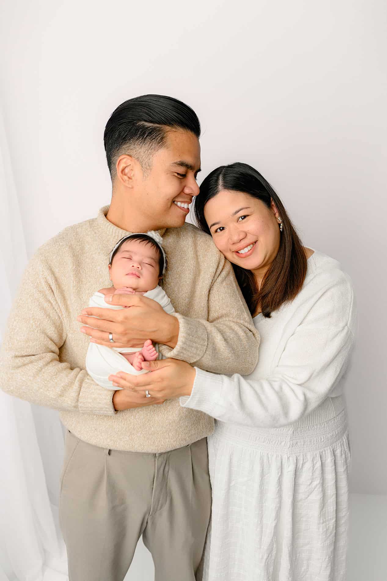 parents standing together holding their newborn baby girl in a white neutral set up 