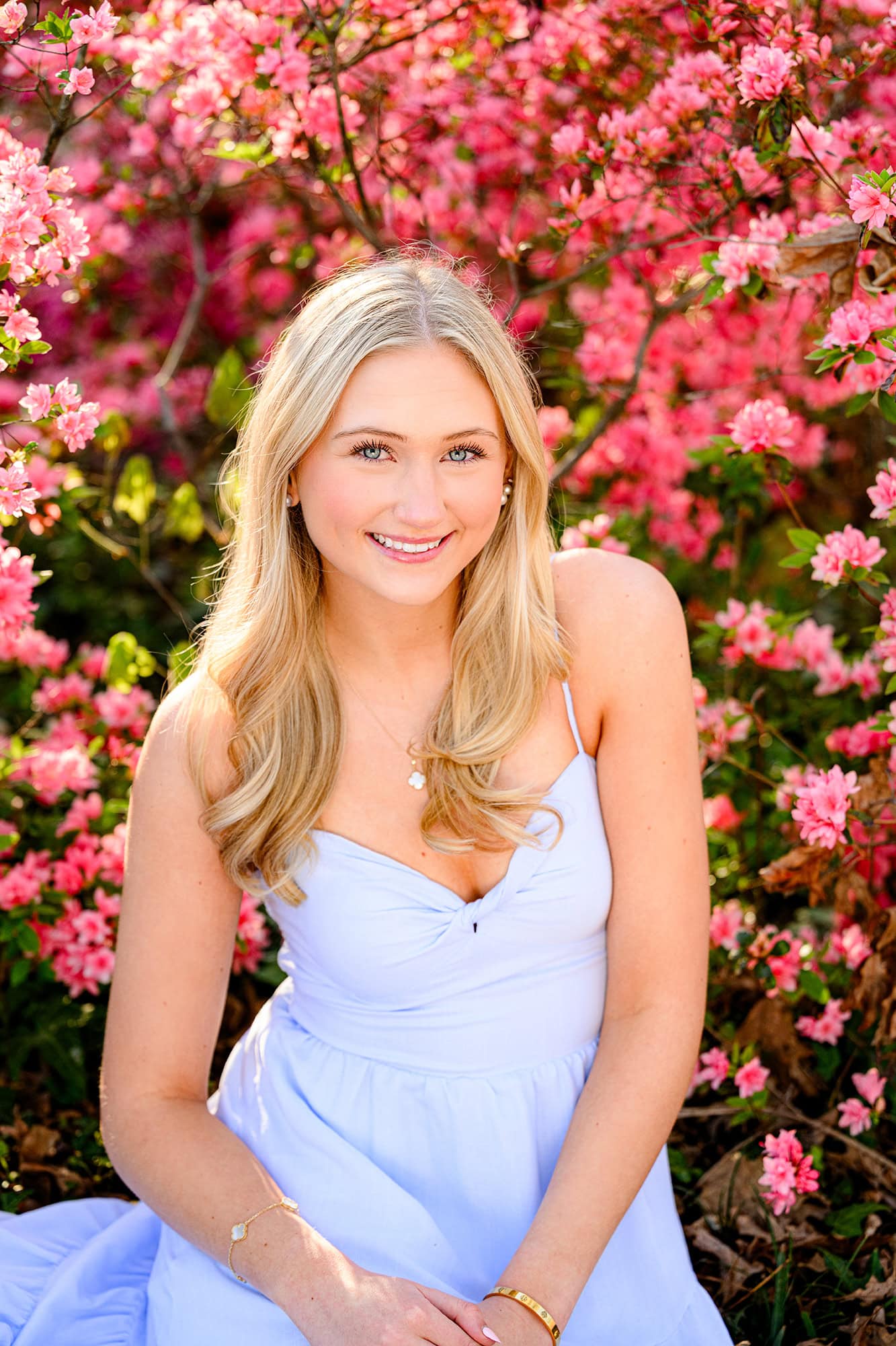 blonde senior sitting in front of pink azaleas wearing a blue dress