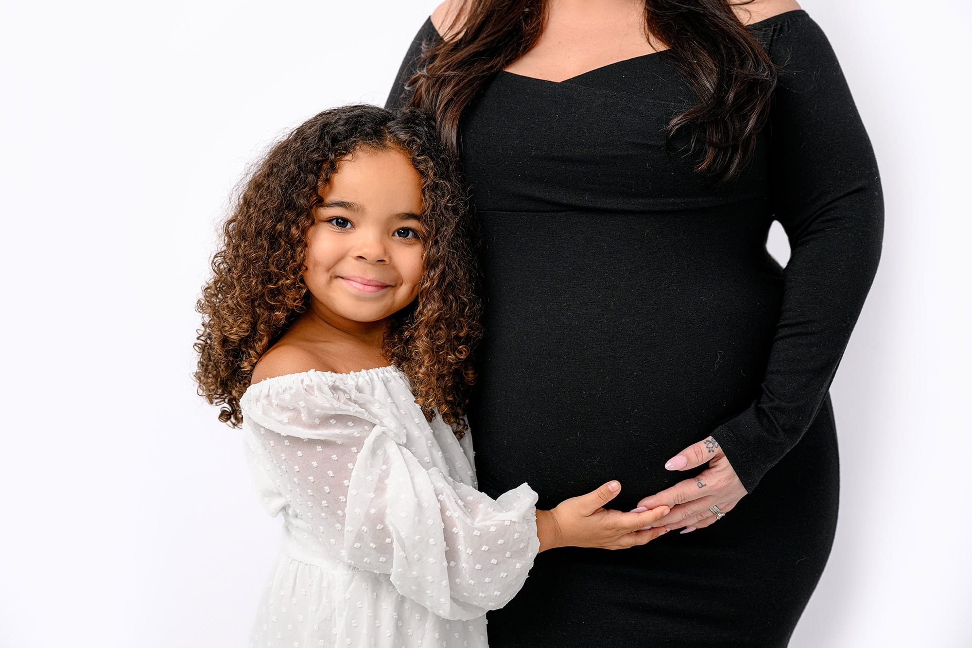 little girl dressed in white standing next to her pregnant mom wearing a black maternity dress holding on to her moms belly
