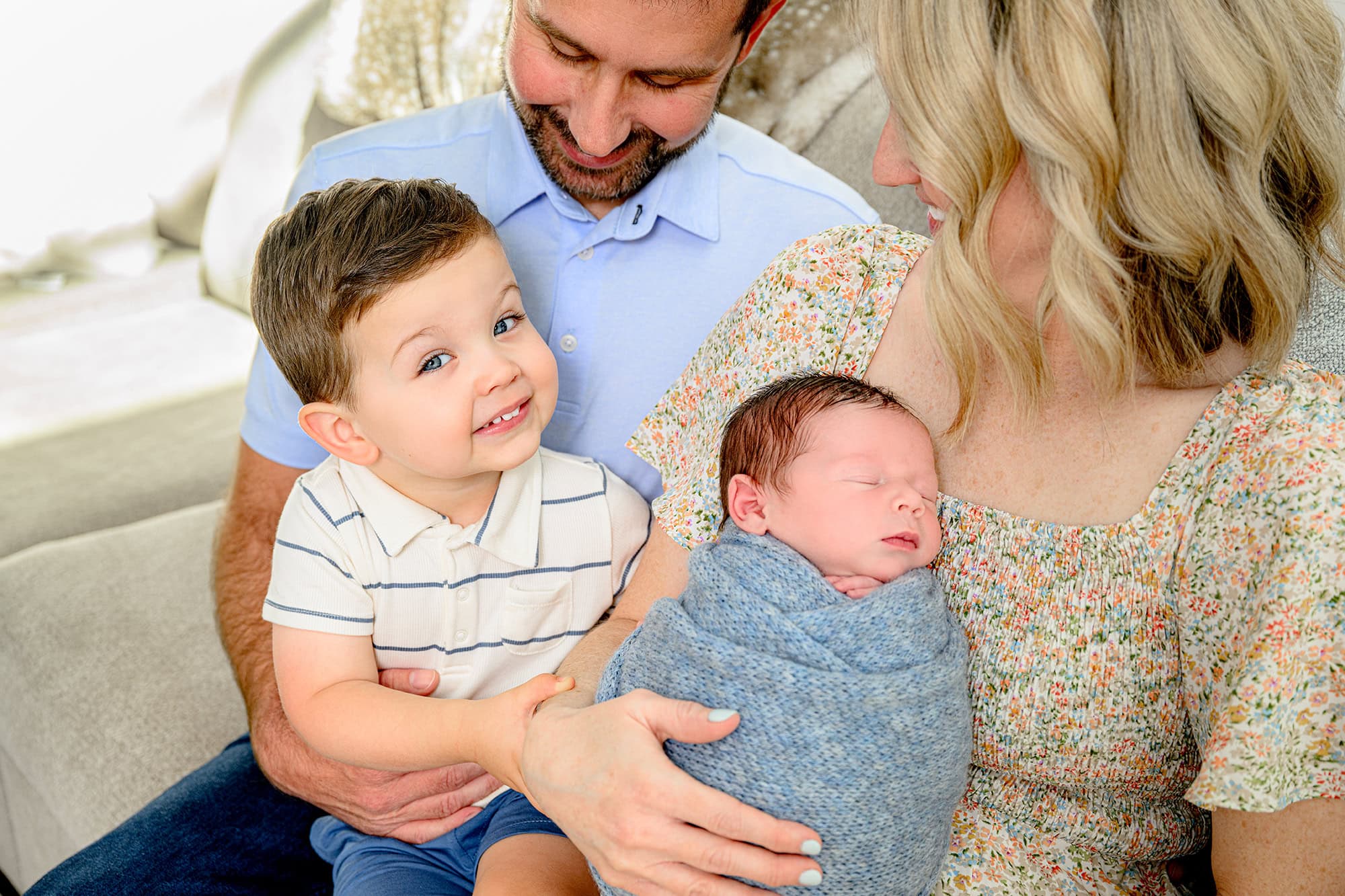 toddler sibling boy grins while he sits in dad's lap during their lifestyle newborn session with mom holding his baby brother
